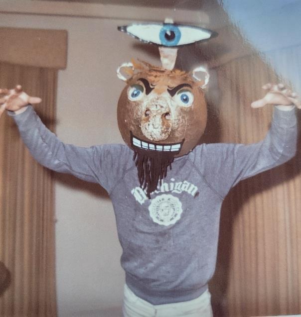 A person stands inside a room wearing a lion mask and a sweatshirt while posing for fun.