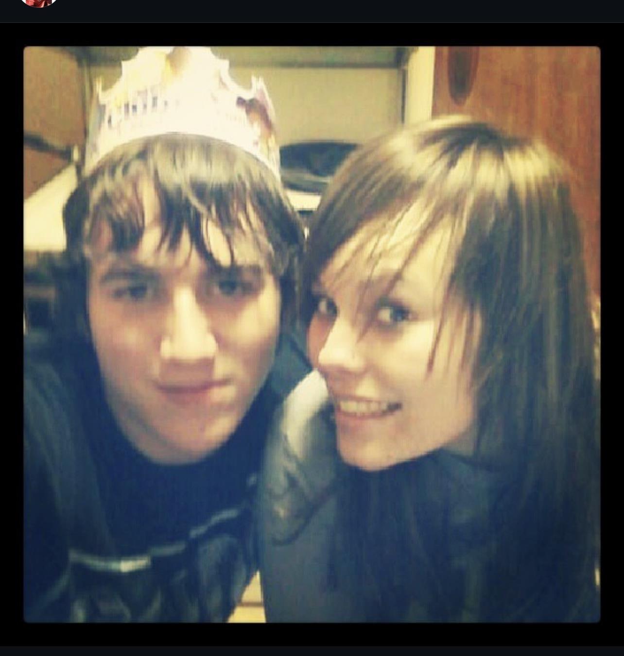 Two young adults pose happily together in a room. The person on the left wears a paper crown.