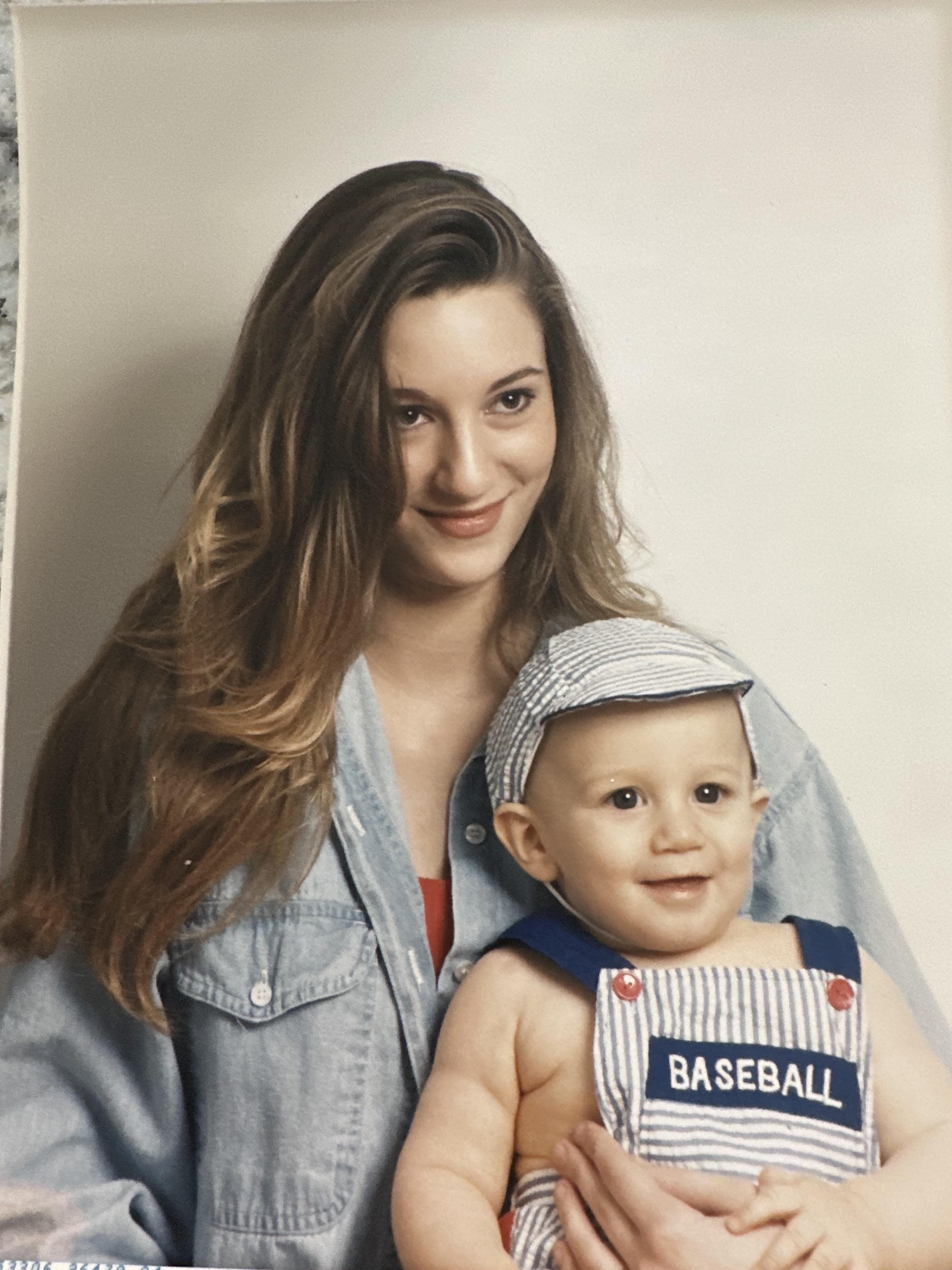 Woman smiles while holding a baby dressed in a baseball outfit inside a building.