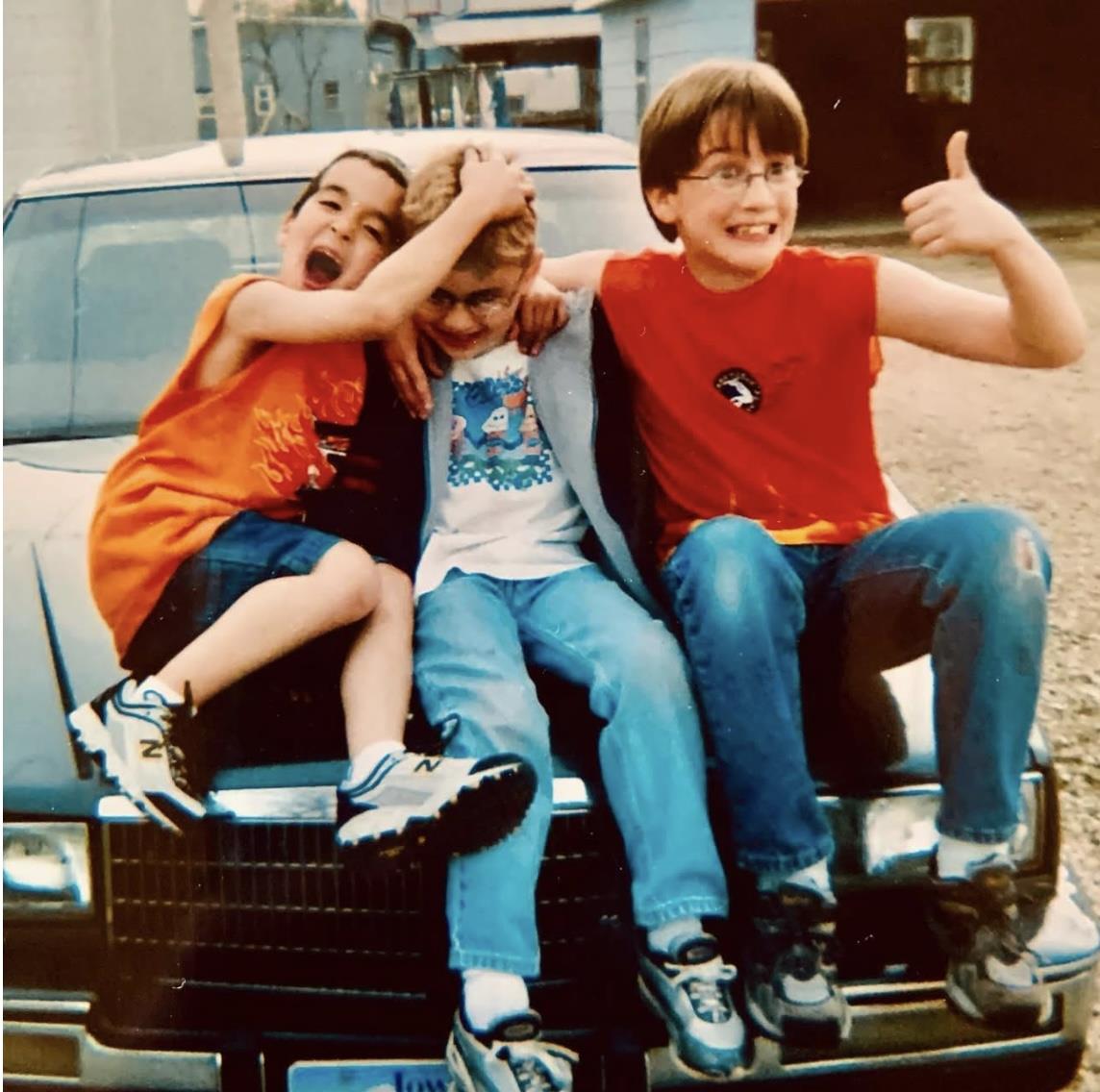 Three kids are on the hood of a car laughing and playing together in a neighborhood setting.