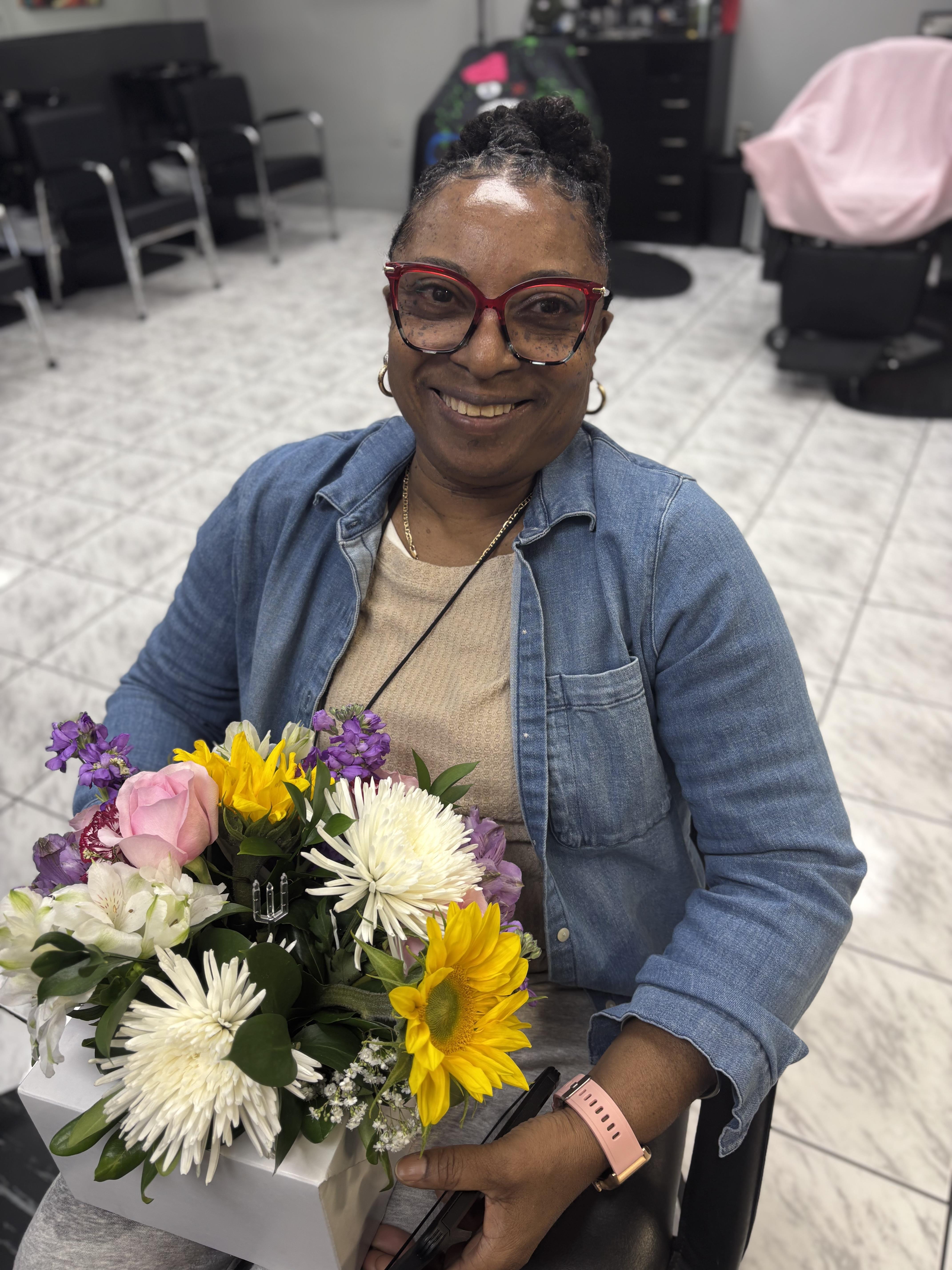 A woman sits in a hair salon holding a colorful flower bouquet while smiling at the camera.