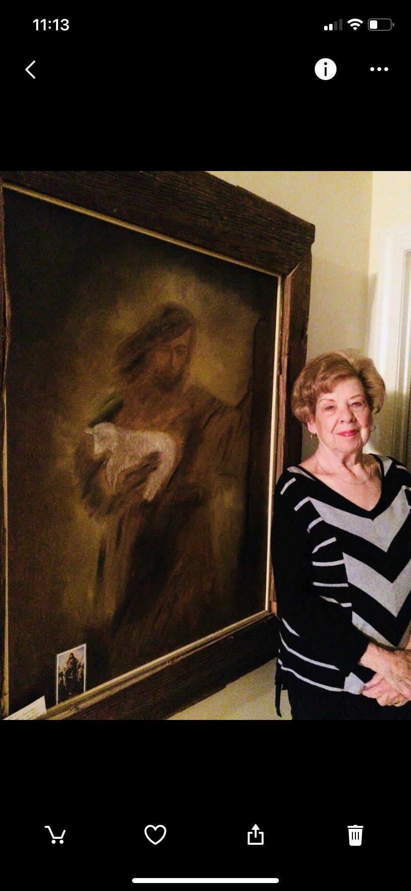 A woman poses beside a large painting of a figure holding a lamb. The setting is an indoor space.