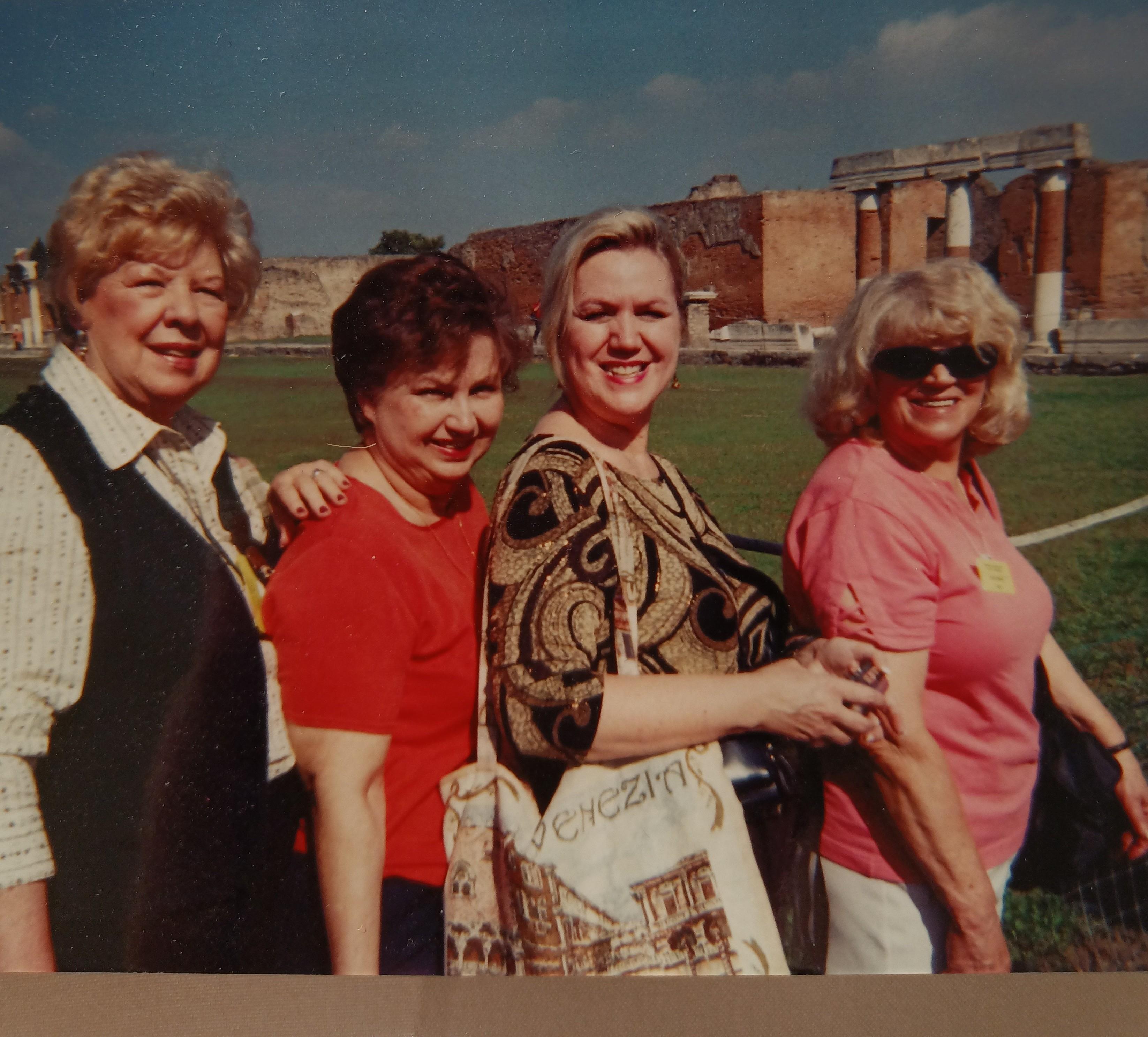 Four women smile and walk side by side on a grassy area with buildings behind them.