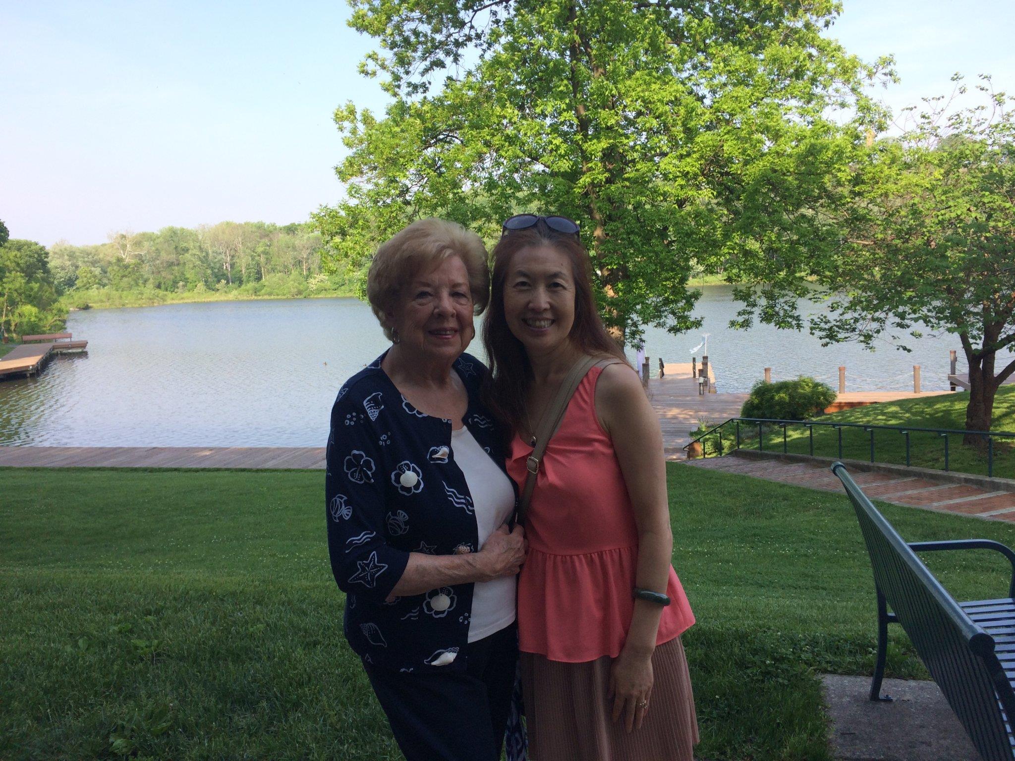 Two people pose for a picture near a lake surrounded by trees on a sunny day.