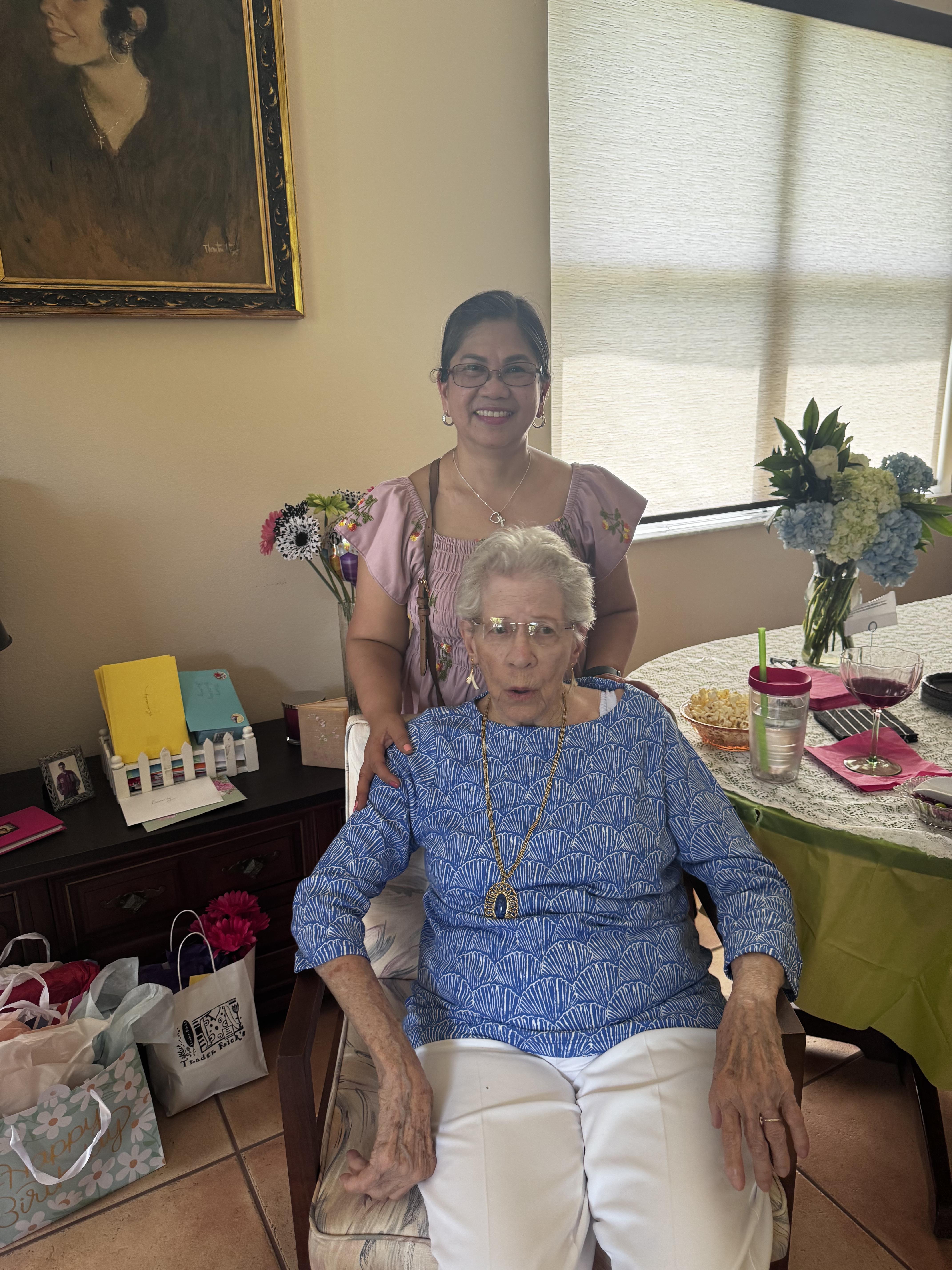 Two women stand and sit together in a living room filled with various items during a gathering.