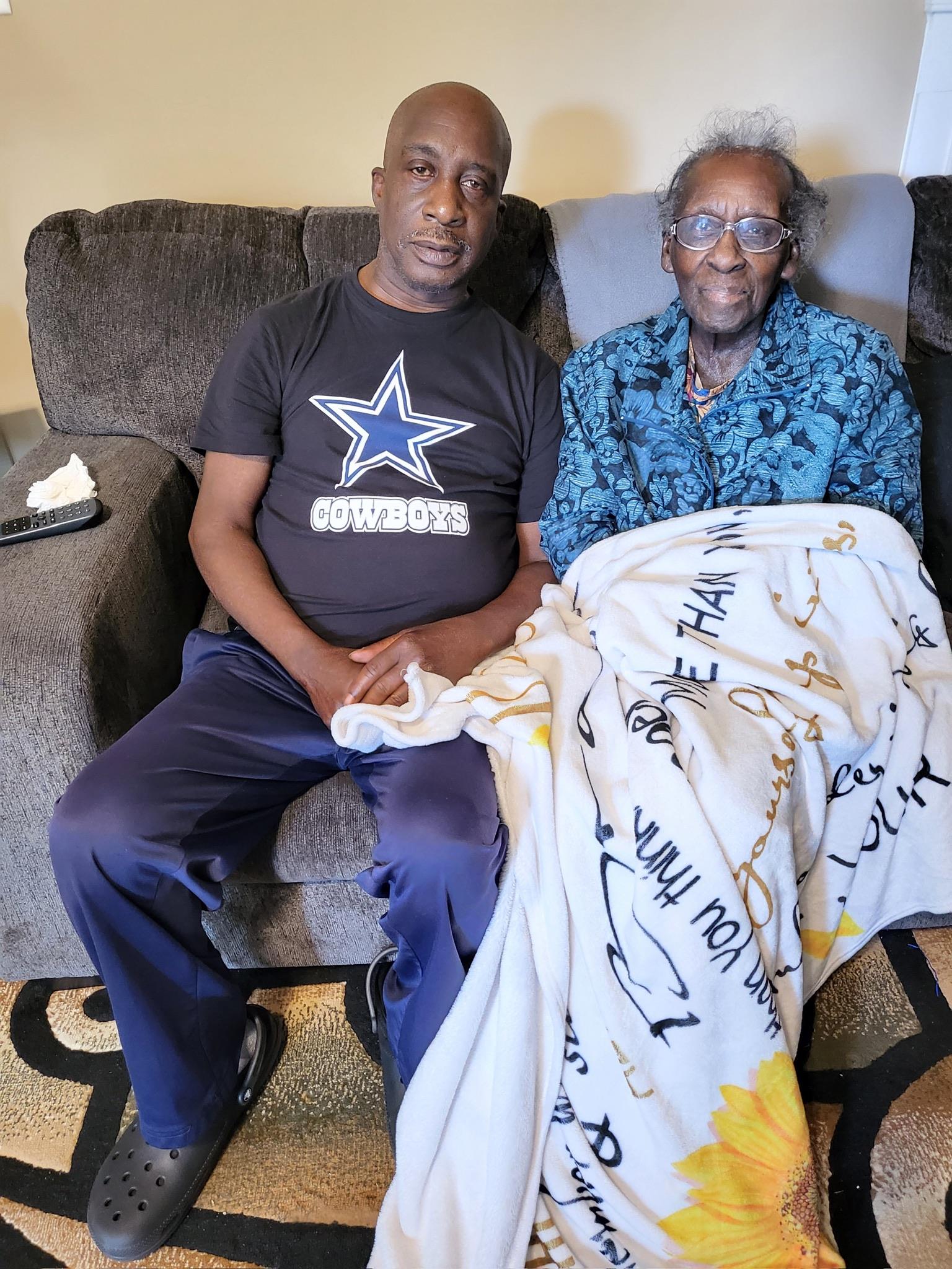 Two men sit together on a couch in a living room, one holds a blanket while they talk.
