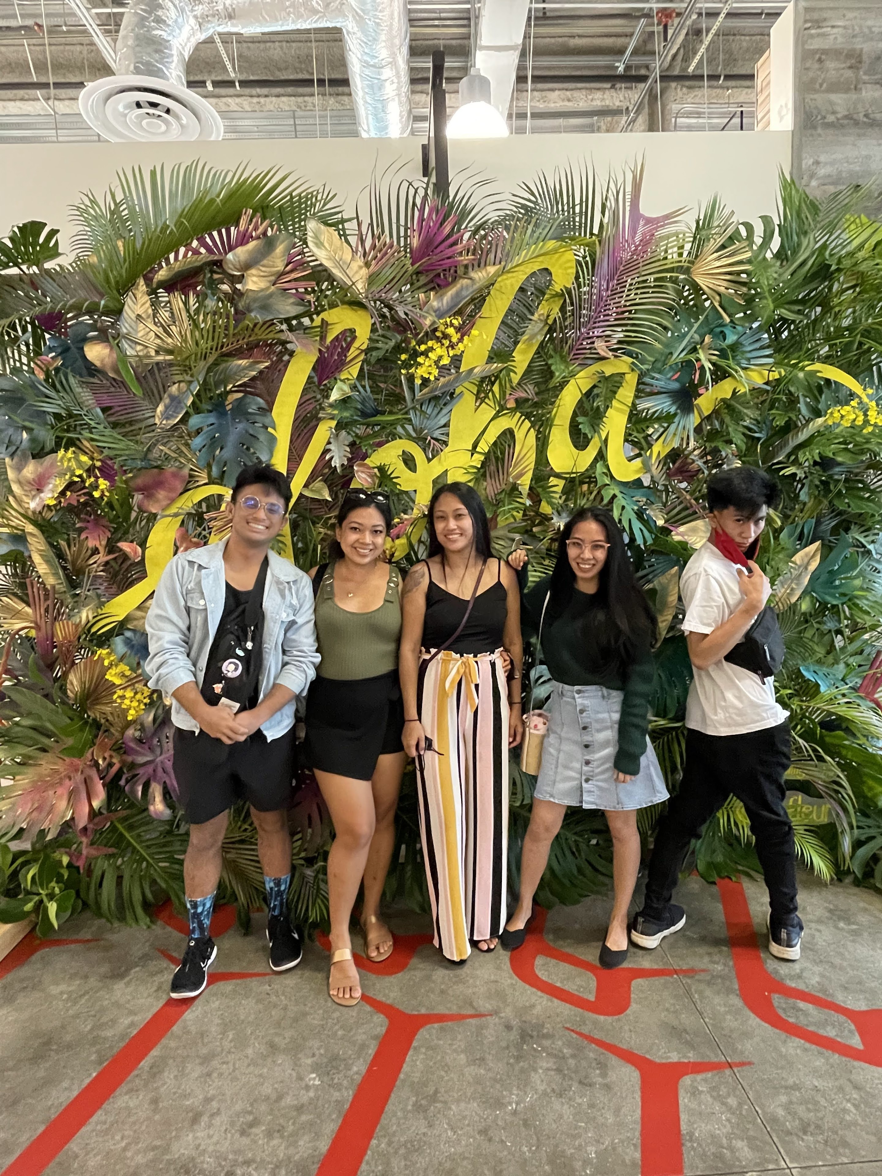 Five friends stand together smiling in front of a vibrant flower wall at a party.