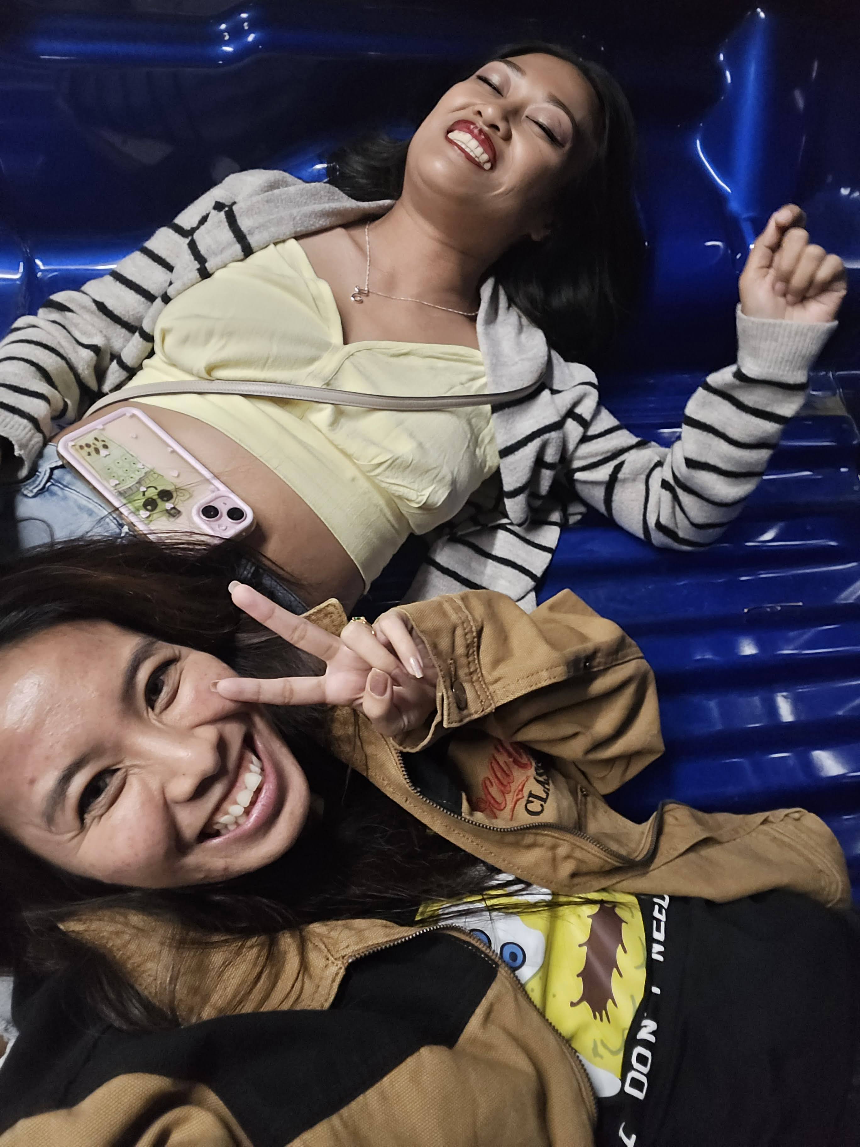Two friends smile and pose while lying on a blue surface, enjoying their time together.