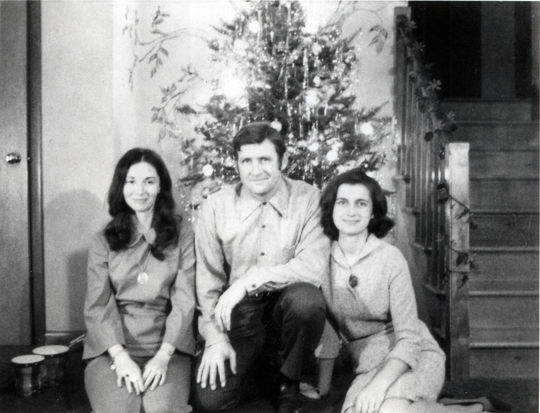Three people gather around a Christmas tree in a cozy home environment during the holidays.