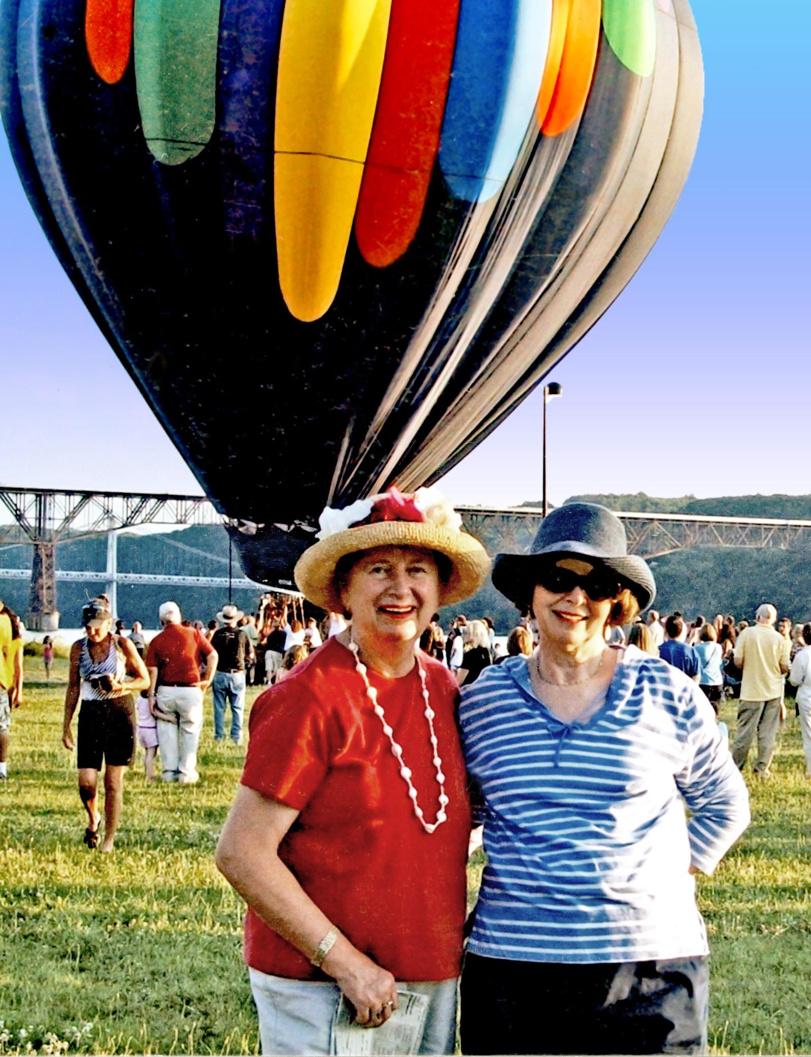 Two women pose happily with a large hot air balloon behind them at a festival in a park.