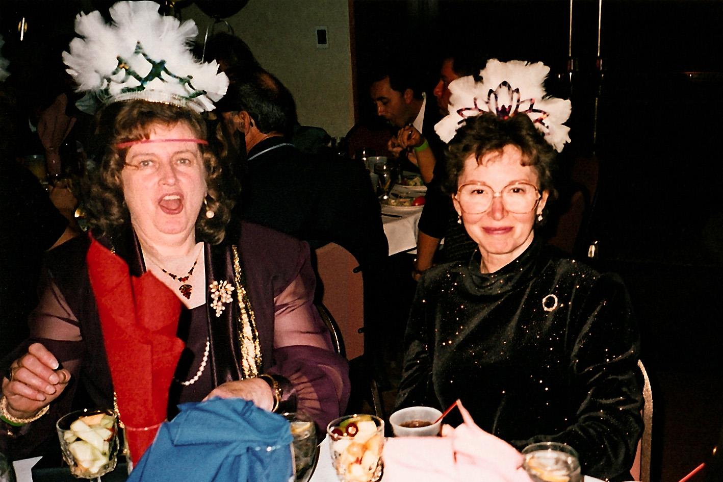 Two women sit together at a table with drinks and food. They wear fancy clothing and headpieces.