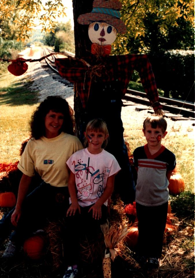 Children and an adult smile together at a pumpkin patch with pumpkins and a scarecrow nearby.