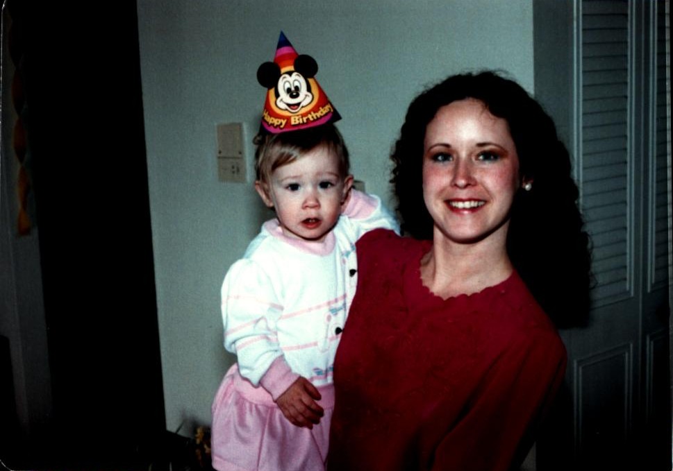 A mother holds her daughter, who wears a birthday hat, while they celebrate a birthday at home.