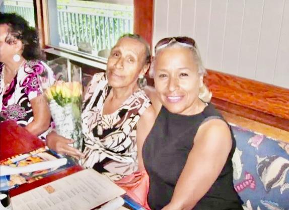 Two women sit together at a table, one holds a bouquet of flowers while both smile.
