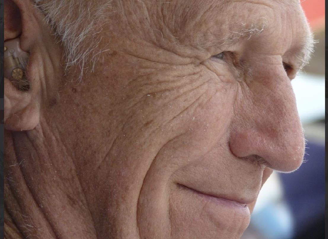 An elderly man sits outdoors with a warm smile on his face. The sunlight highlights his features.