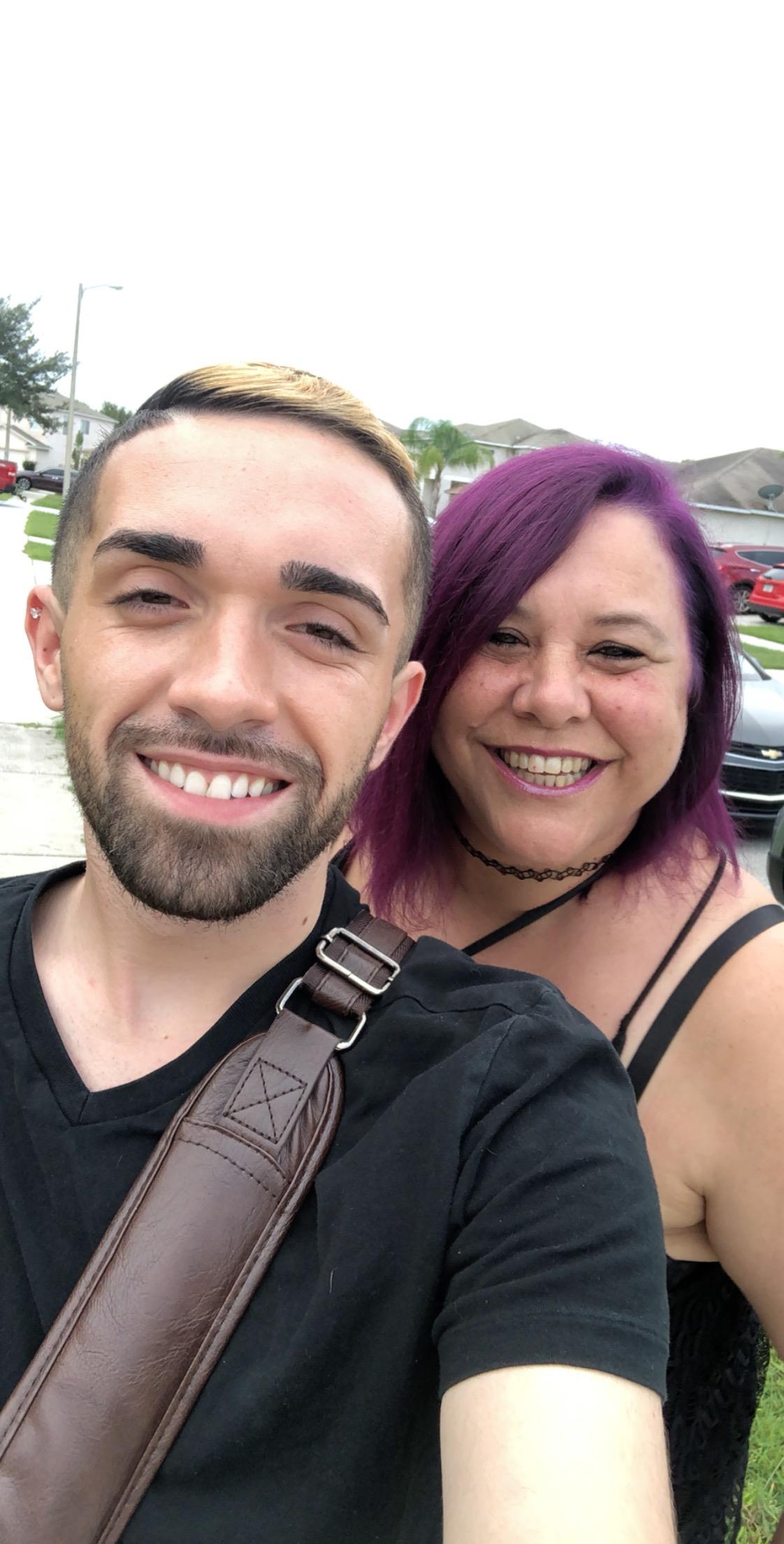 Two people smile as they take a selfie outside in a neighborhood with houses nearby.