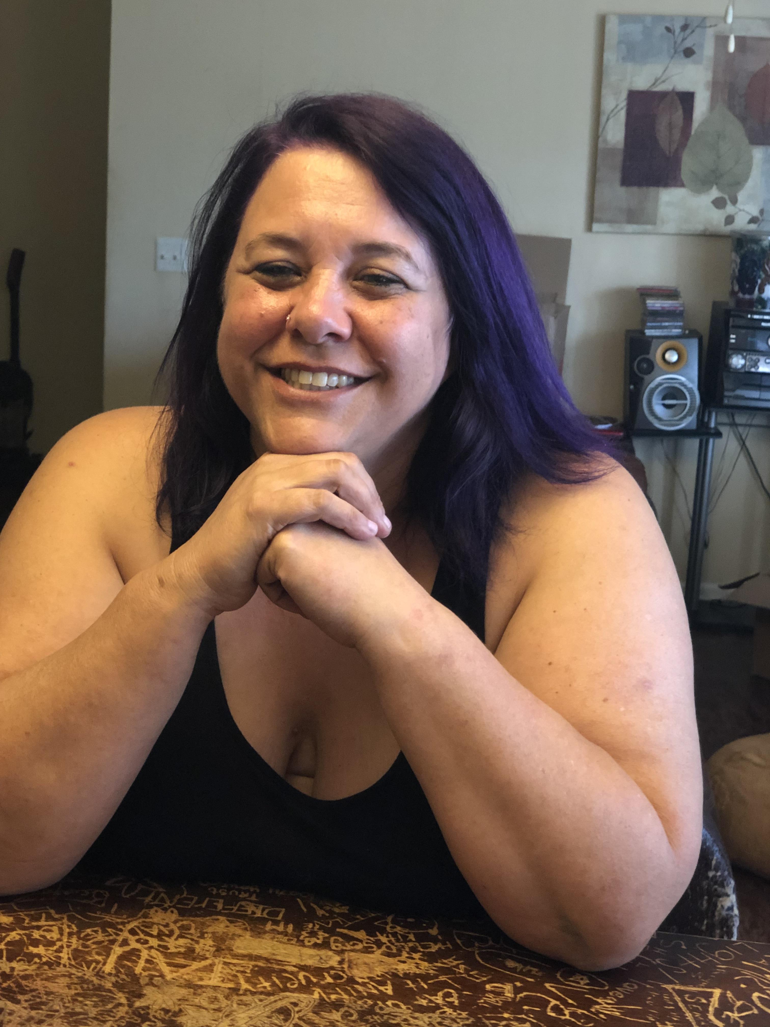 A woman is smiling at the camera while resting her chin on her hands in a home setting.