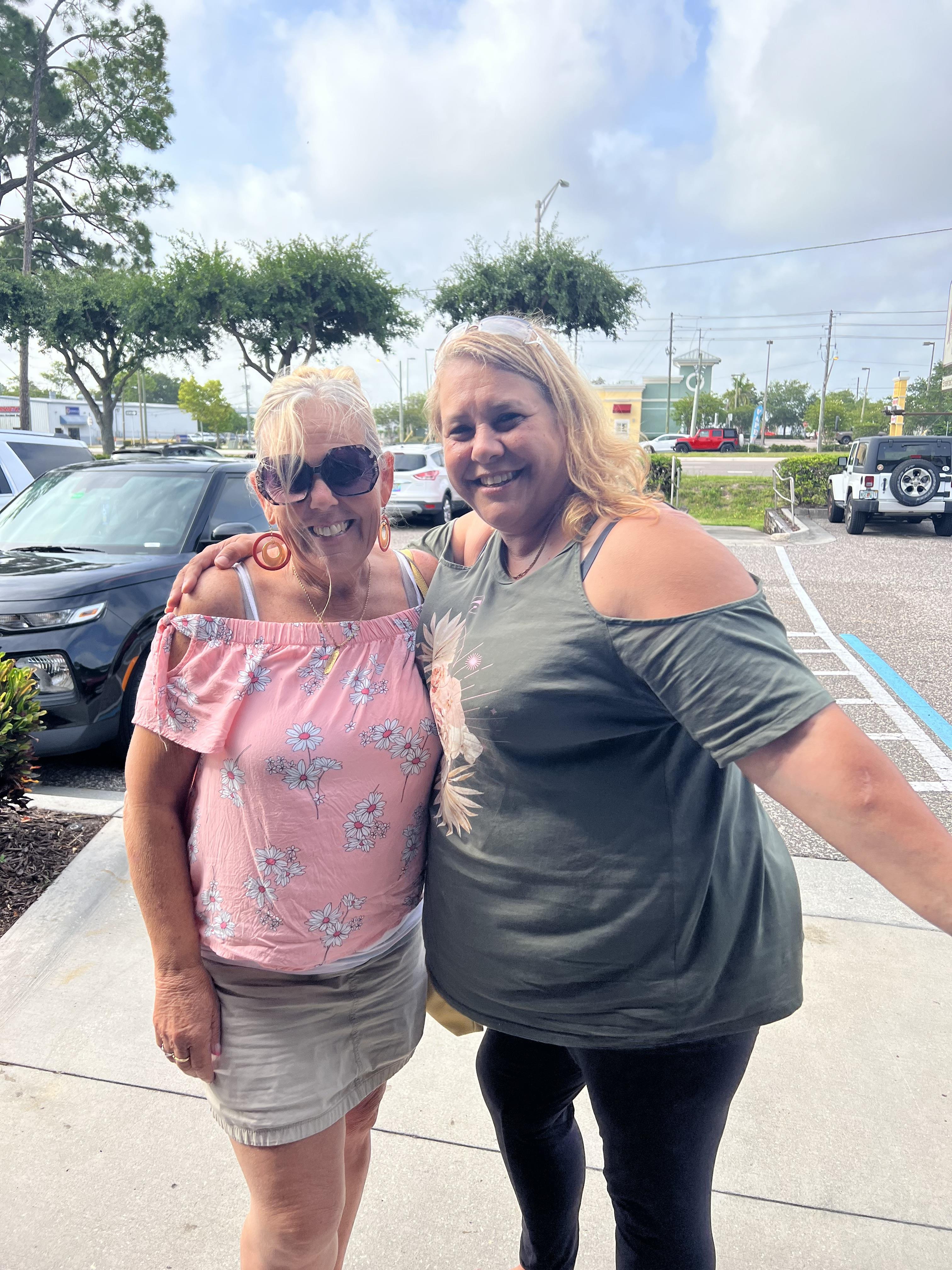 Two women stand together outside a store, smiling and enjoying their time on a sunny day.