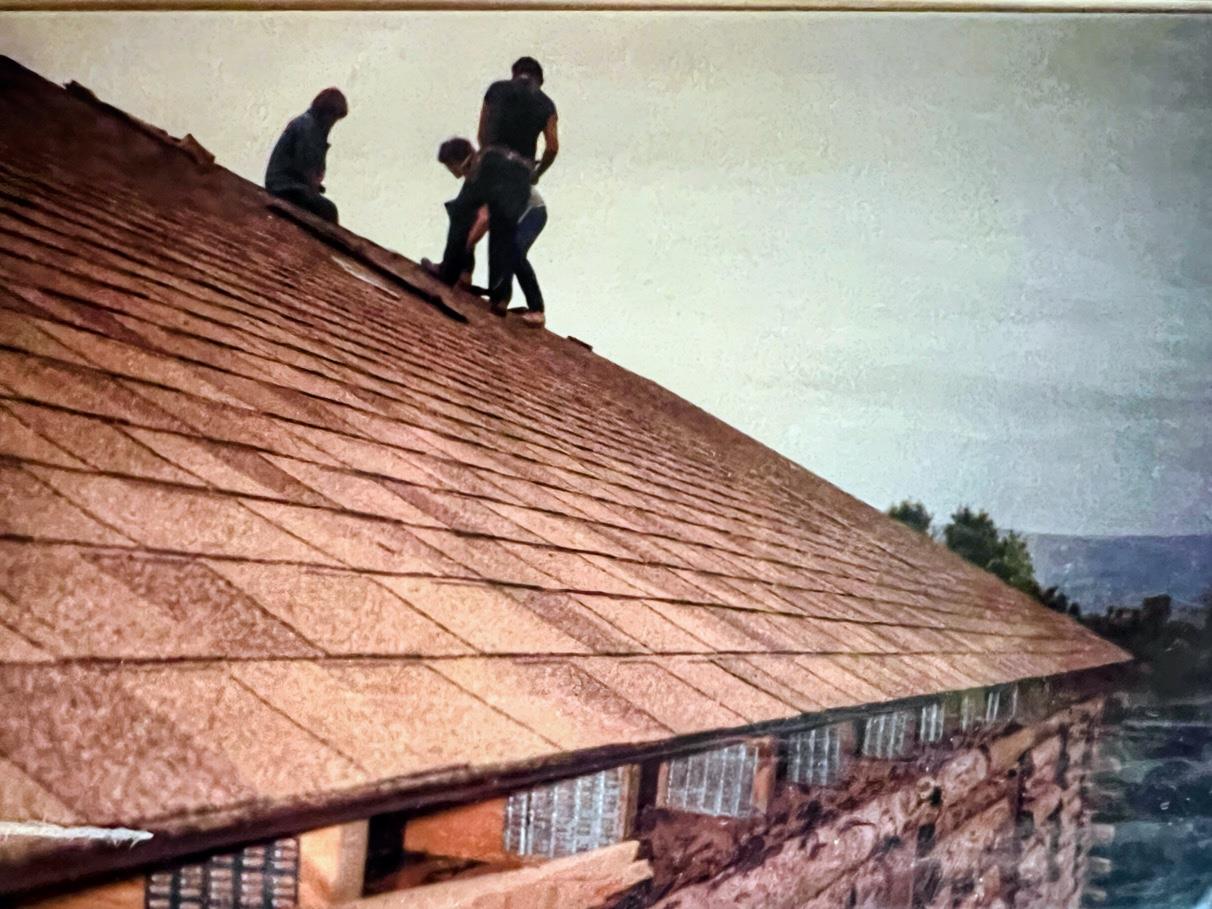 Three workers are on the roof putting shingles while one looks down at the ground.