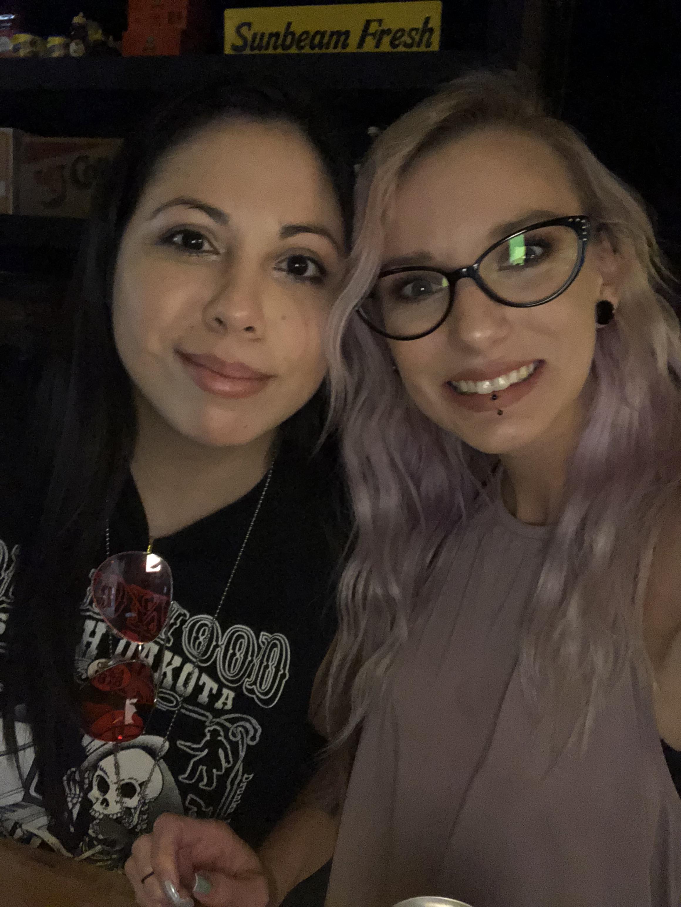 Two women are close together smiling at a social event in a dark setting.