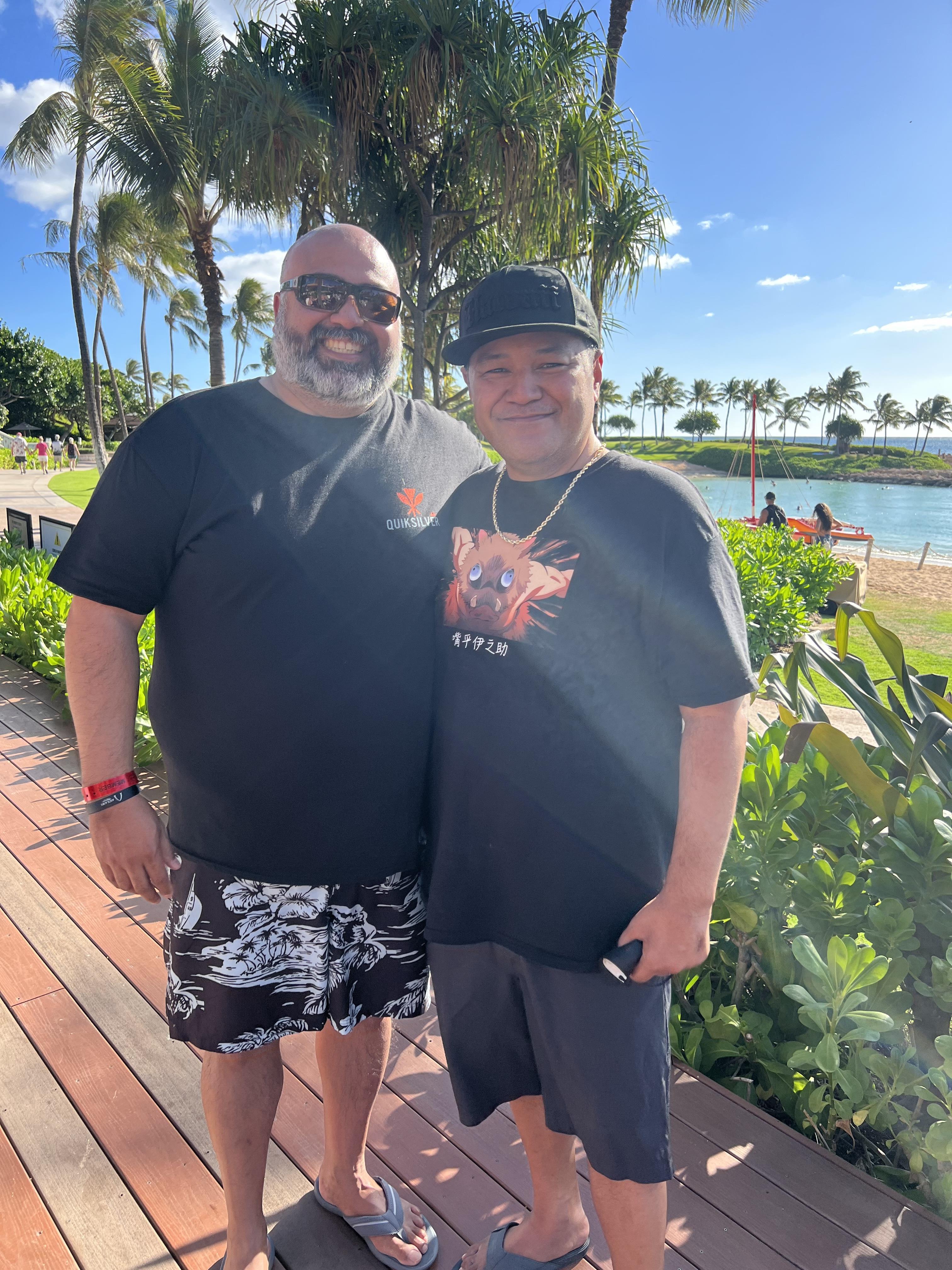 Two men smile and pose together near a beach surrounded by palm trees and blue skies.