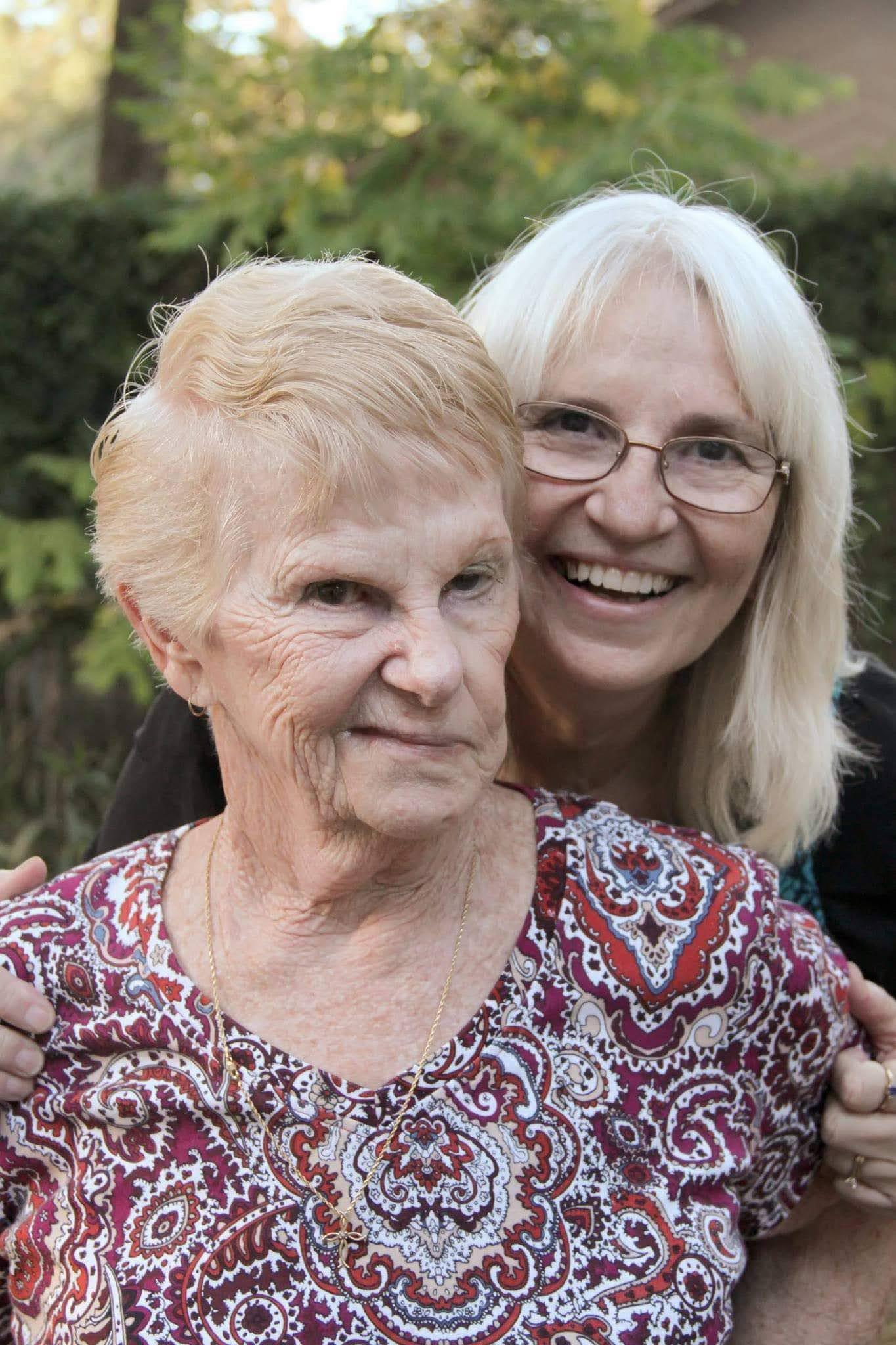 Two women stand close together with smiles on their faces, enjoying time outdoors in a park.