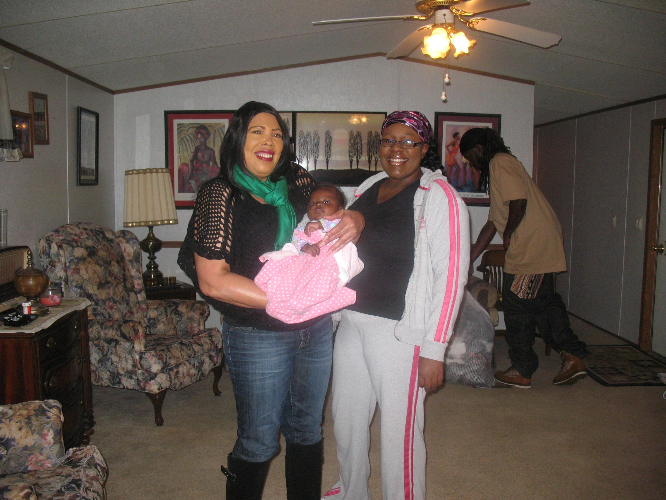 Two women stand with a baby in a living room while another person organizes items in the background.