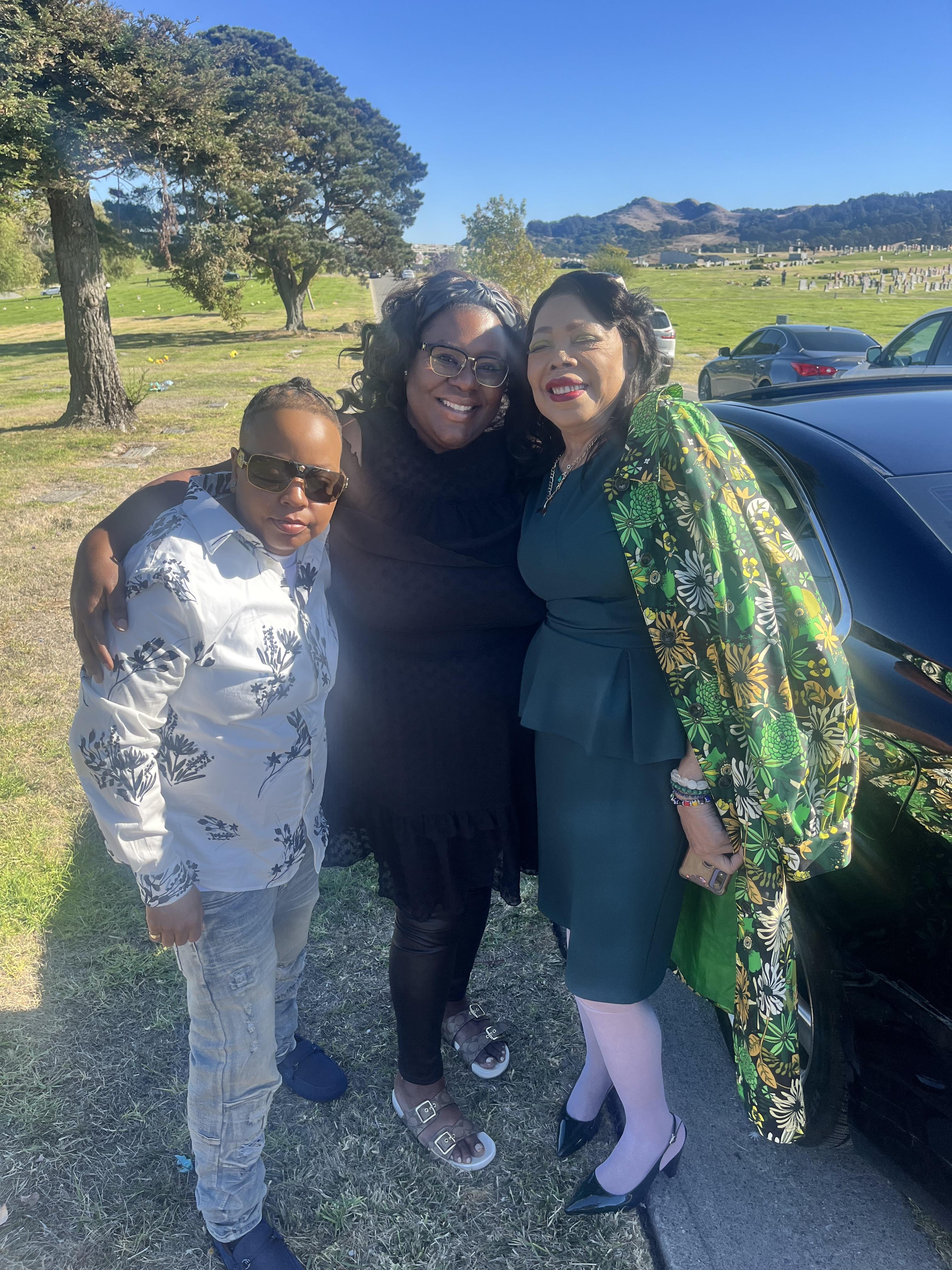 Three people stand close to a car in a park with green grass and trees.