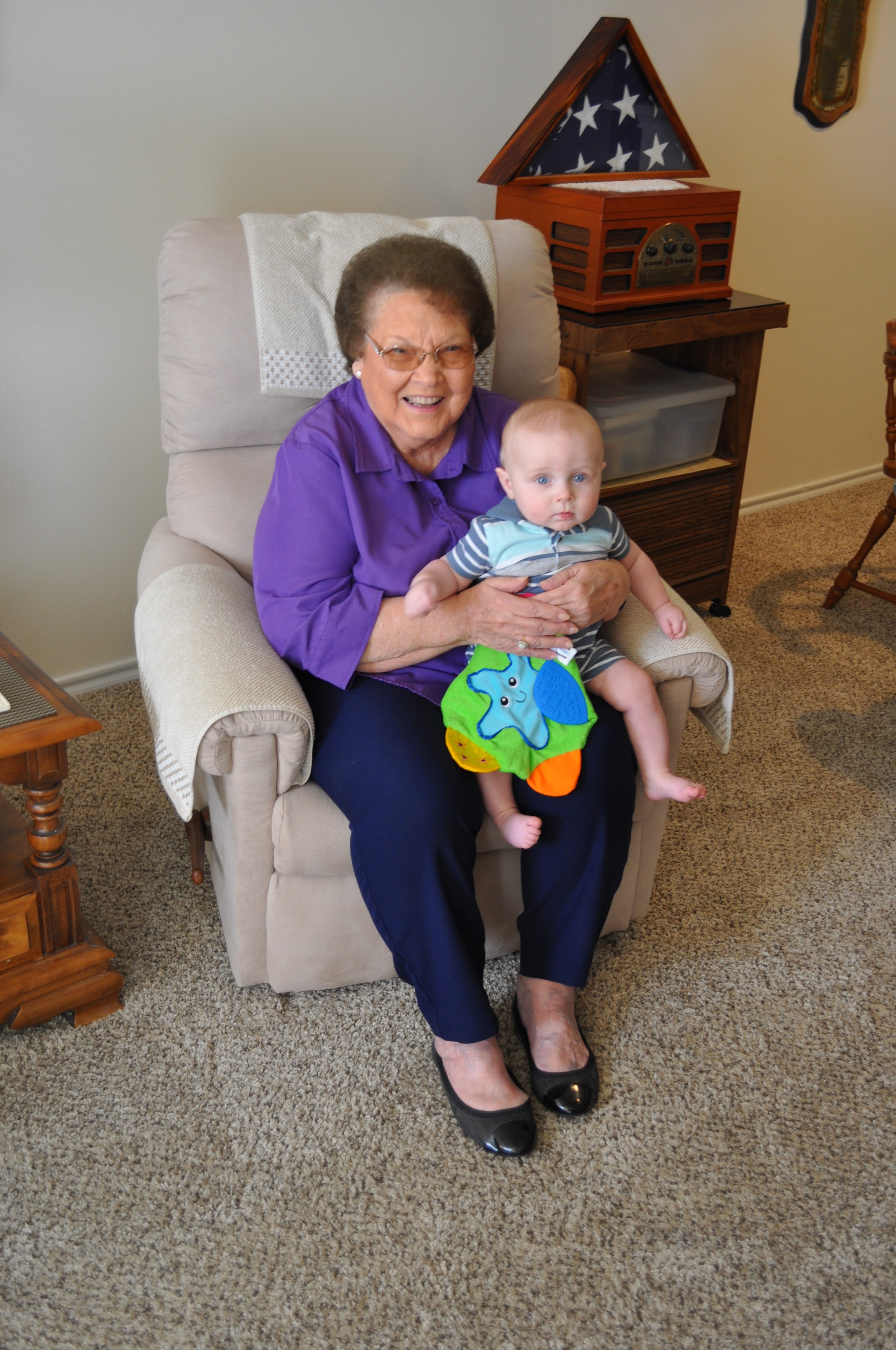 An elderly woman with glasses smiles while holding a baby on her lap in a chair indoors.