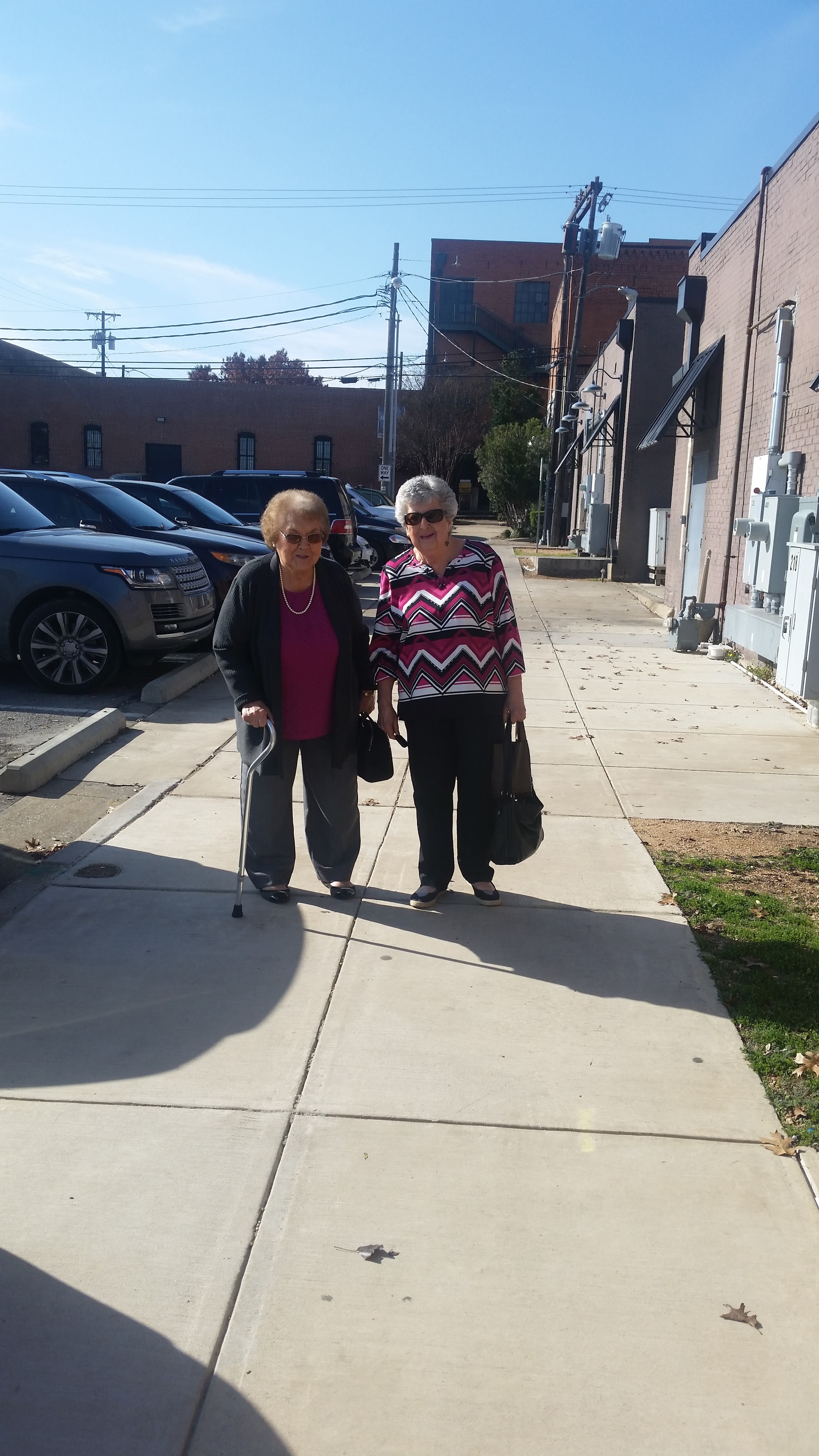 Two elder women walk side by side on the sidewalk. They are near parked cars in a city.