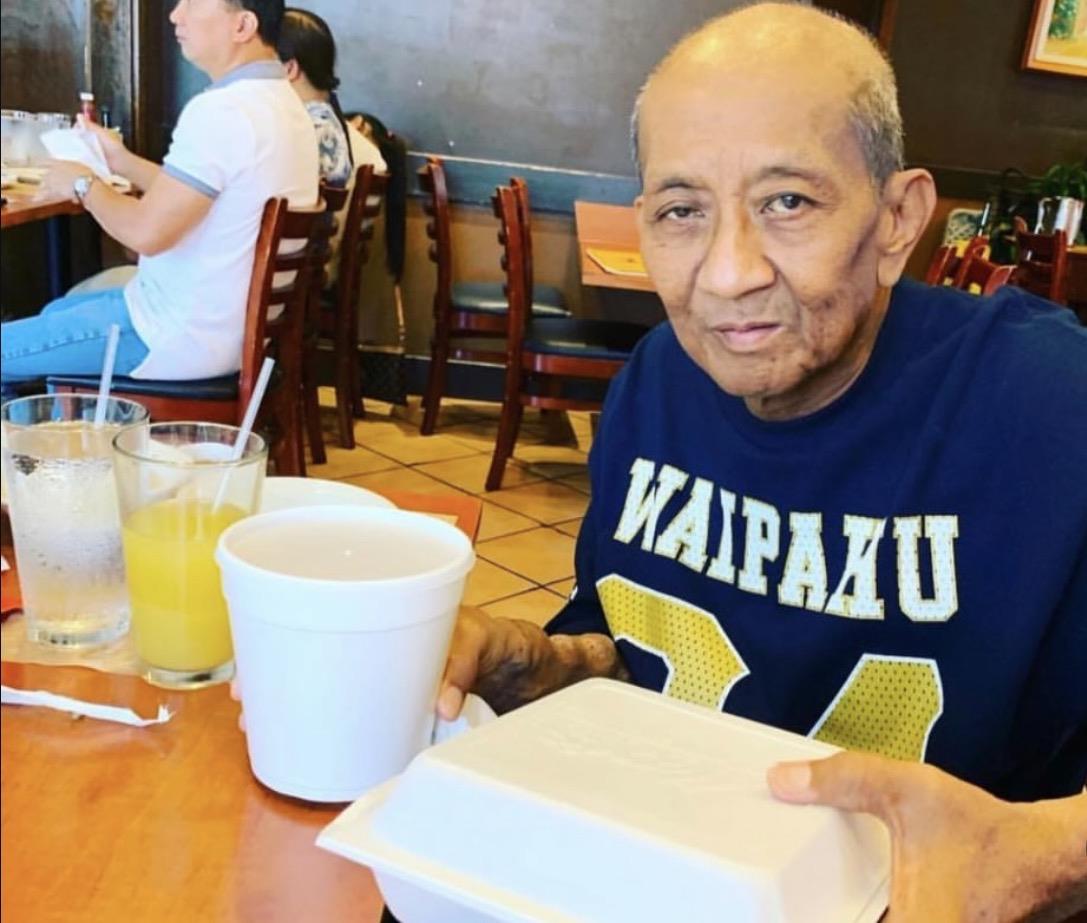 An elderly man sits at a restaurant table holding a food box. Friends talk in the background.
