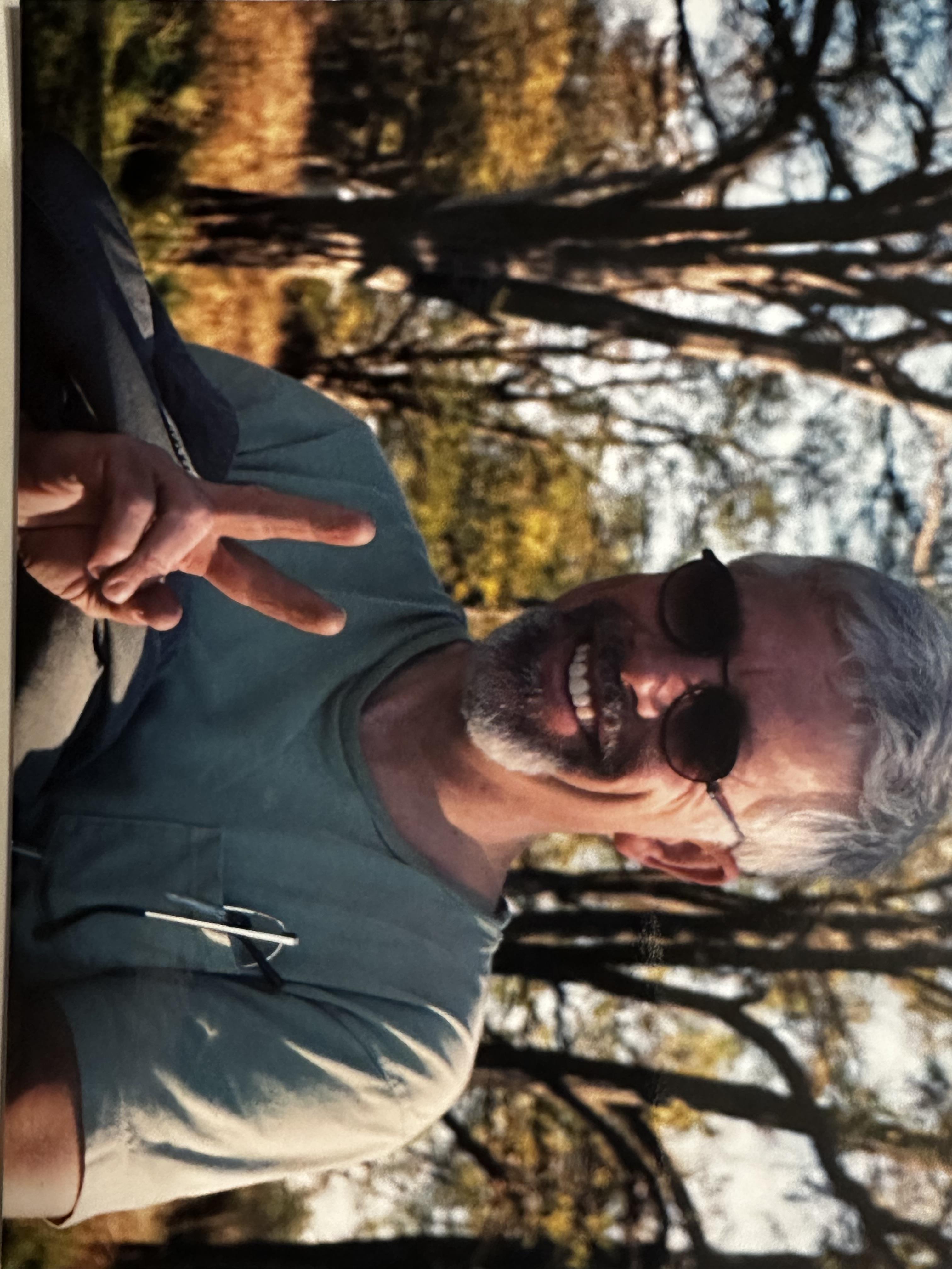 A man smiles and gives a peace sign while sitting outdoors in a natural environment with trees.