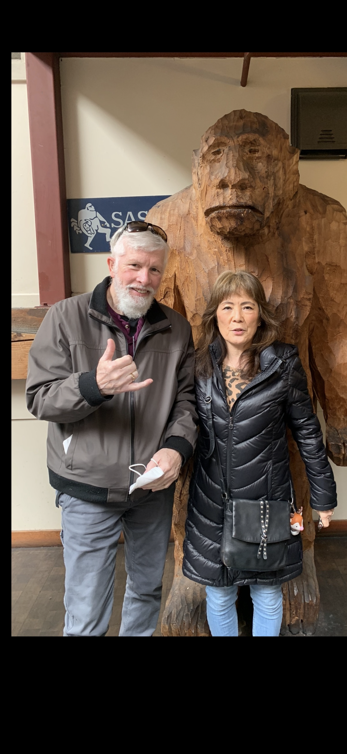 A man and a woman stand beside a large wooden bear statue while smiling for a picture.