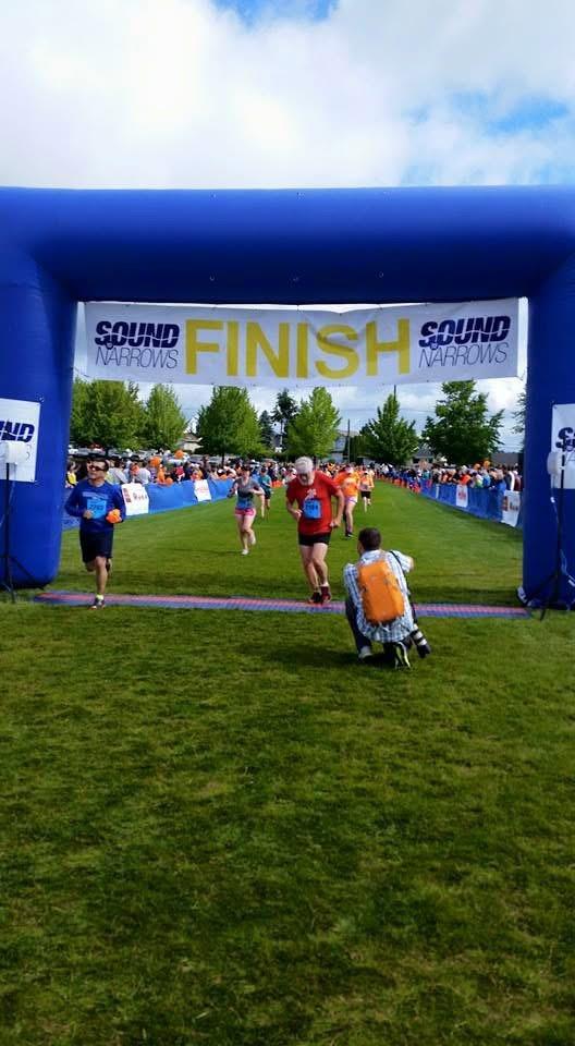 Participants run toward the finish line while spectators cheer and take photos in a park setting.