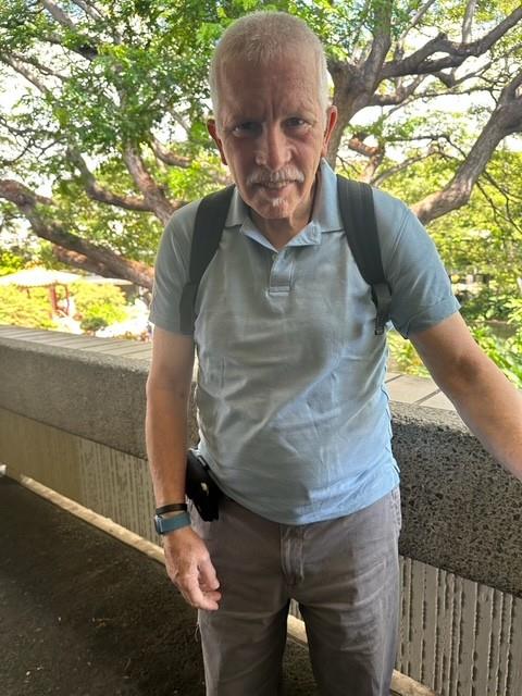 A senior man stands at a terrace with trees and greenery in the background.