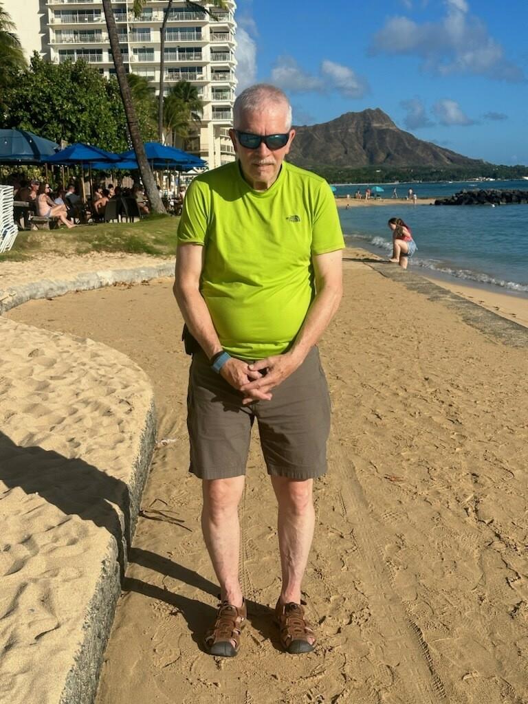 An elderly man stands on the beach with water and a famous mountain behind him in the sun.