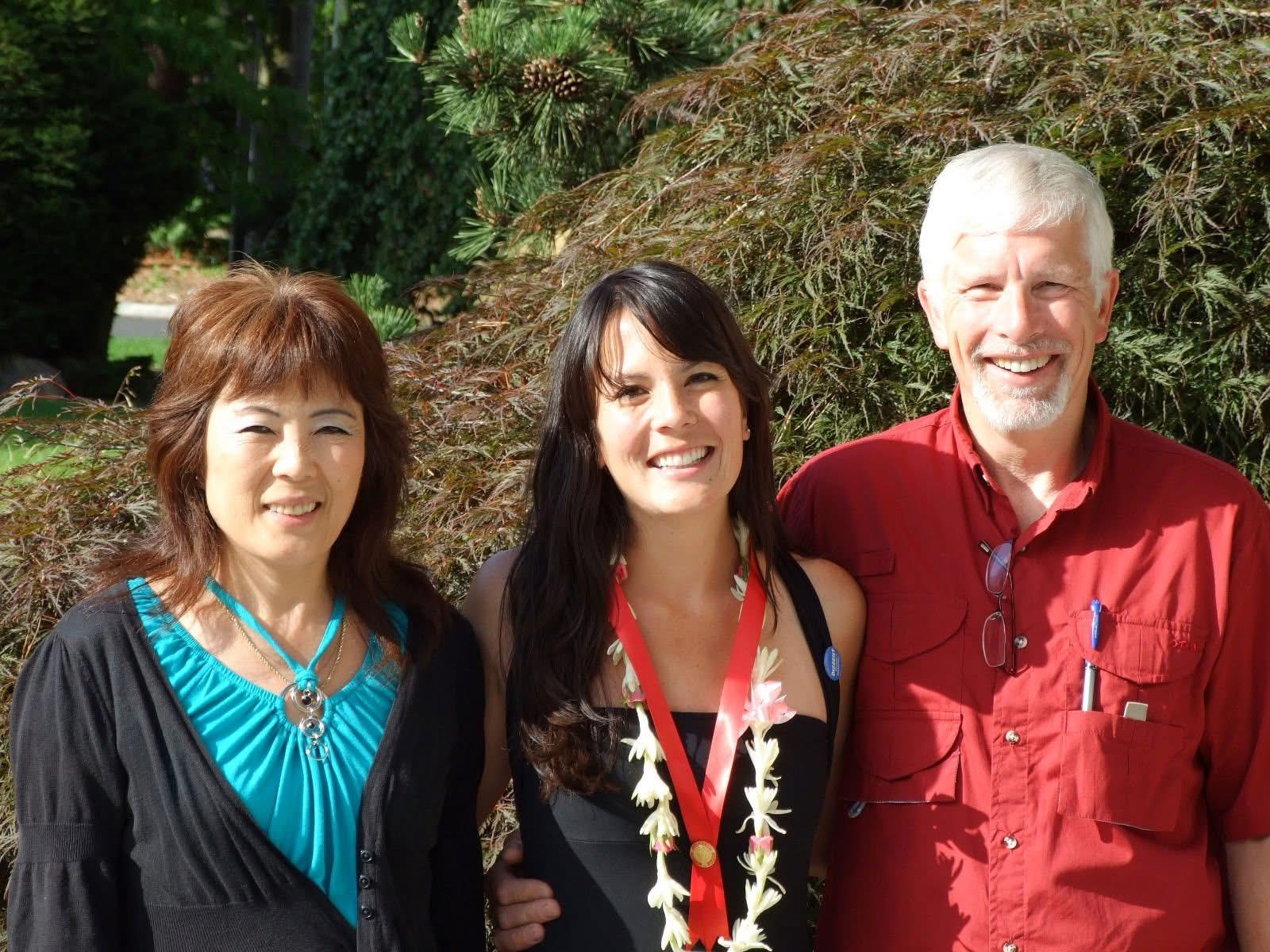 Three friends smile together outdoors in a garden with greenery and sunlight around them.