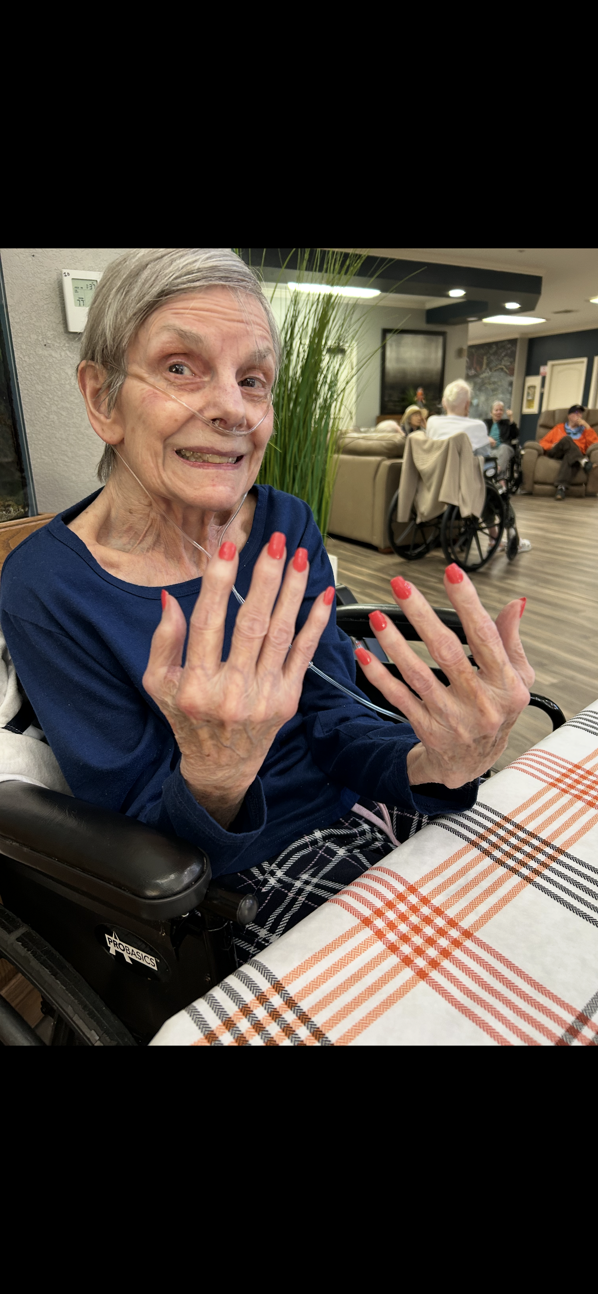 An elderly woman smiles and displays her painted nails while sitting in a chair.