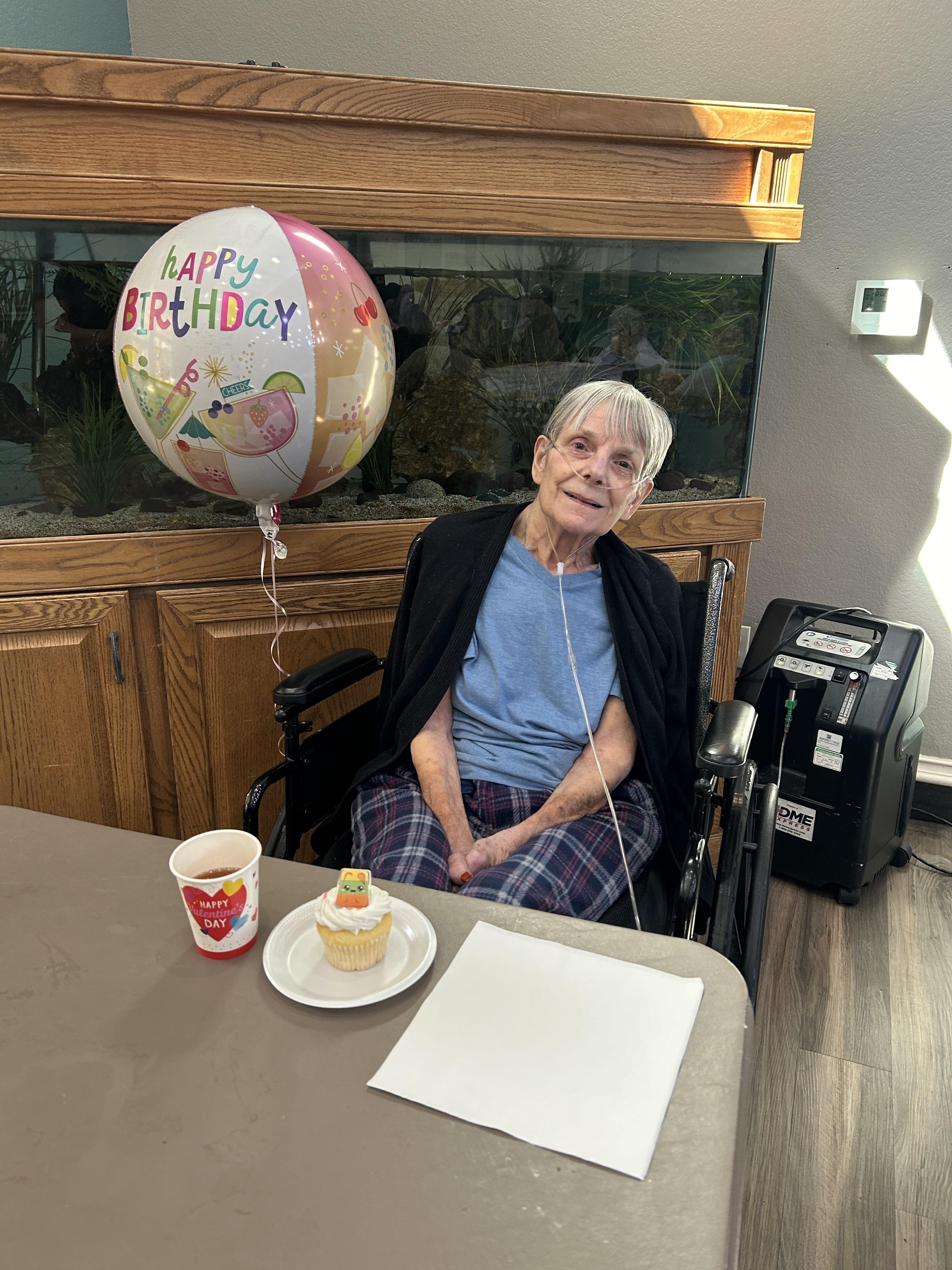 A senior person sits in a wheelchair celebrating their birthday with a cupcake and balloon.