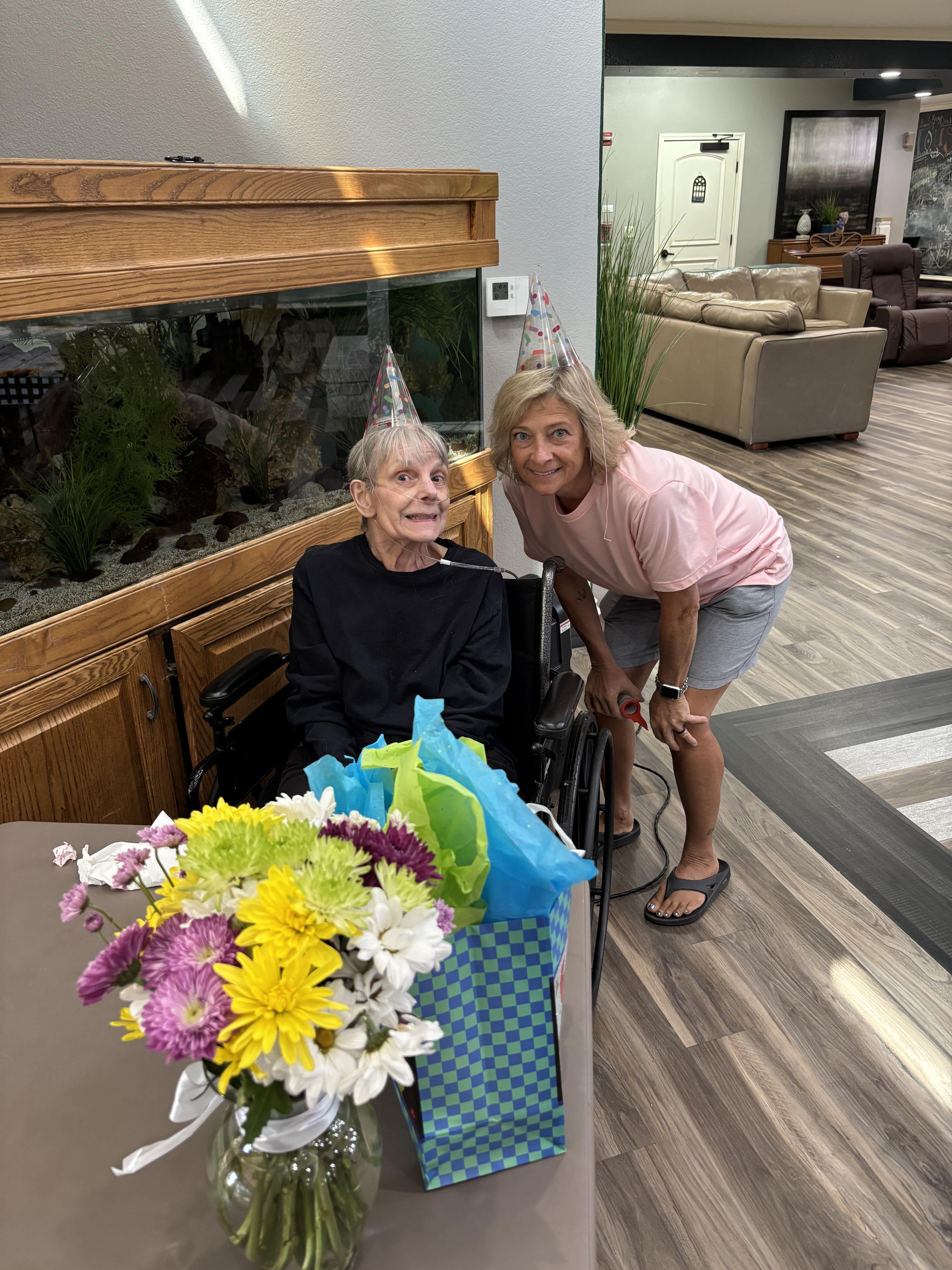 A woman and her friend celebrate a birthday in a bright living room with flowers and gifts.