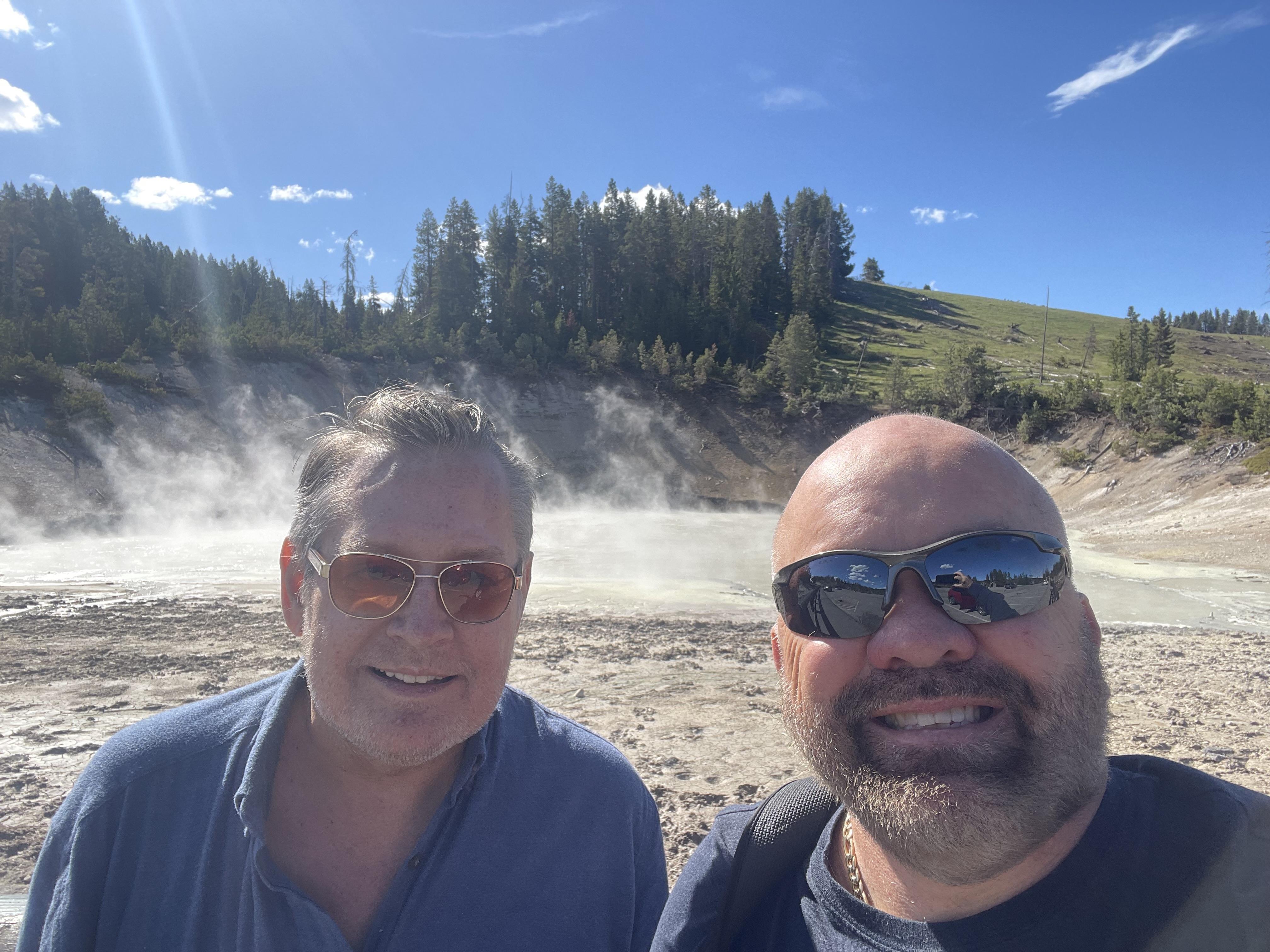 Two friends smile for a selfie at Yellowstone, with geysers and trees in the background.