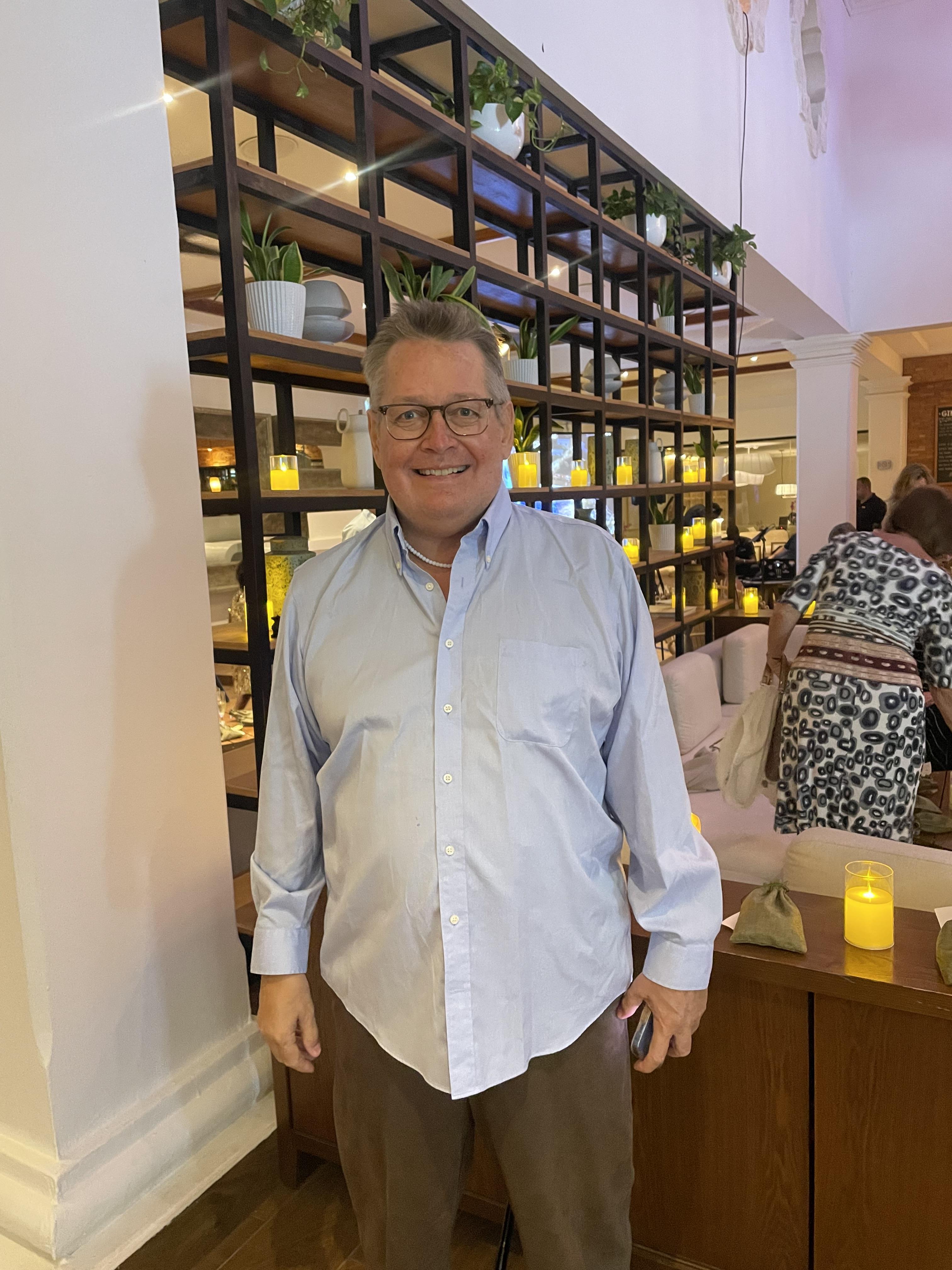 A man stands in a cafe with a bookshelf behind him while people sit and talk.