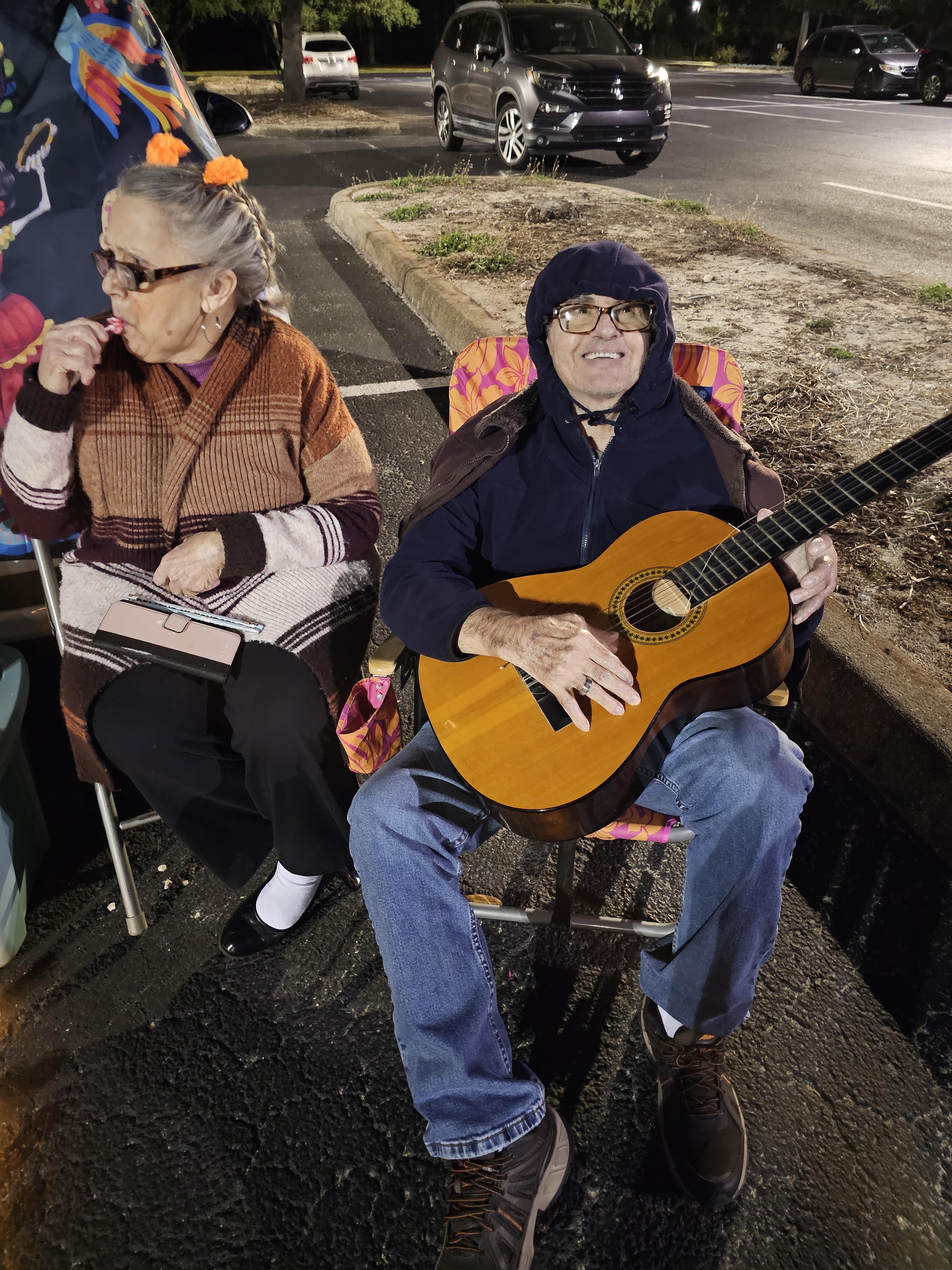 An elderly couple shares snacks while one plays a guitar under street lights at night.