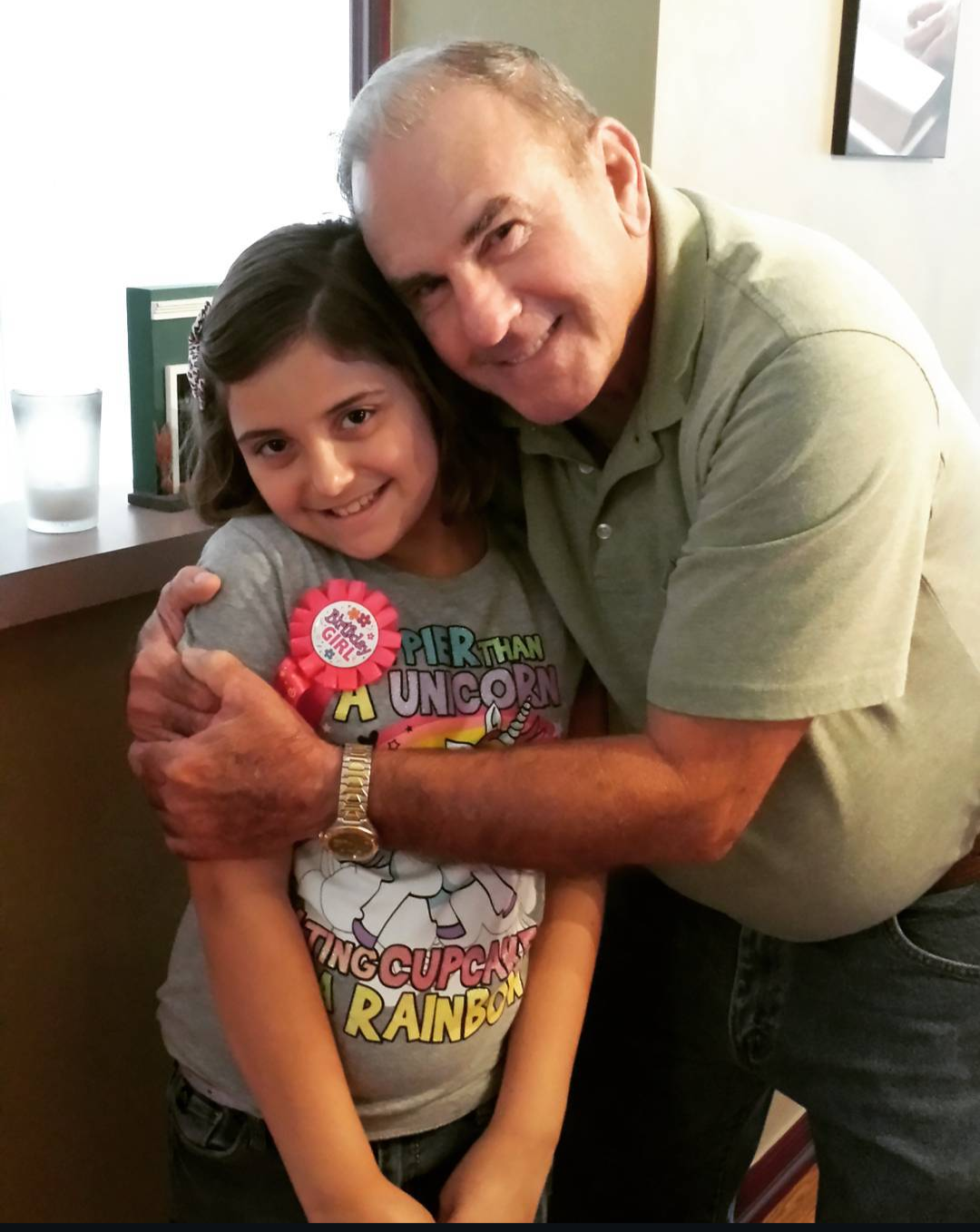 A grandfather hugs his granddaughter while they smile at a birthday gathering in a room.