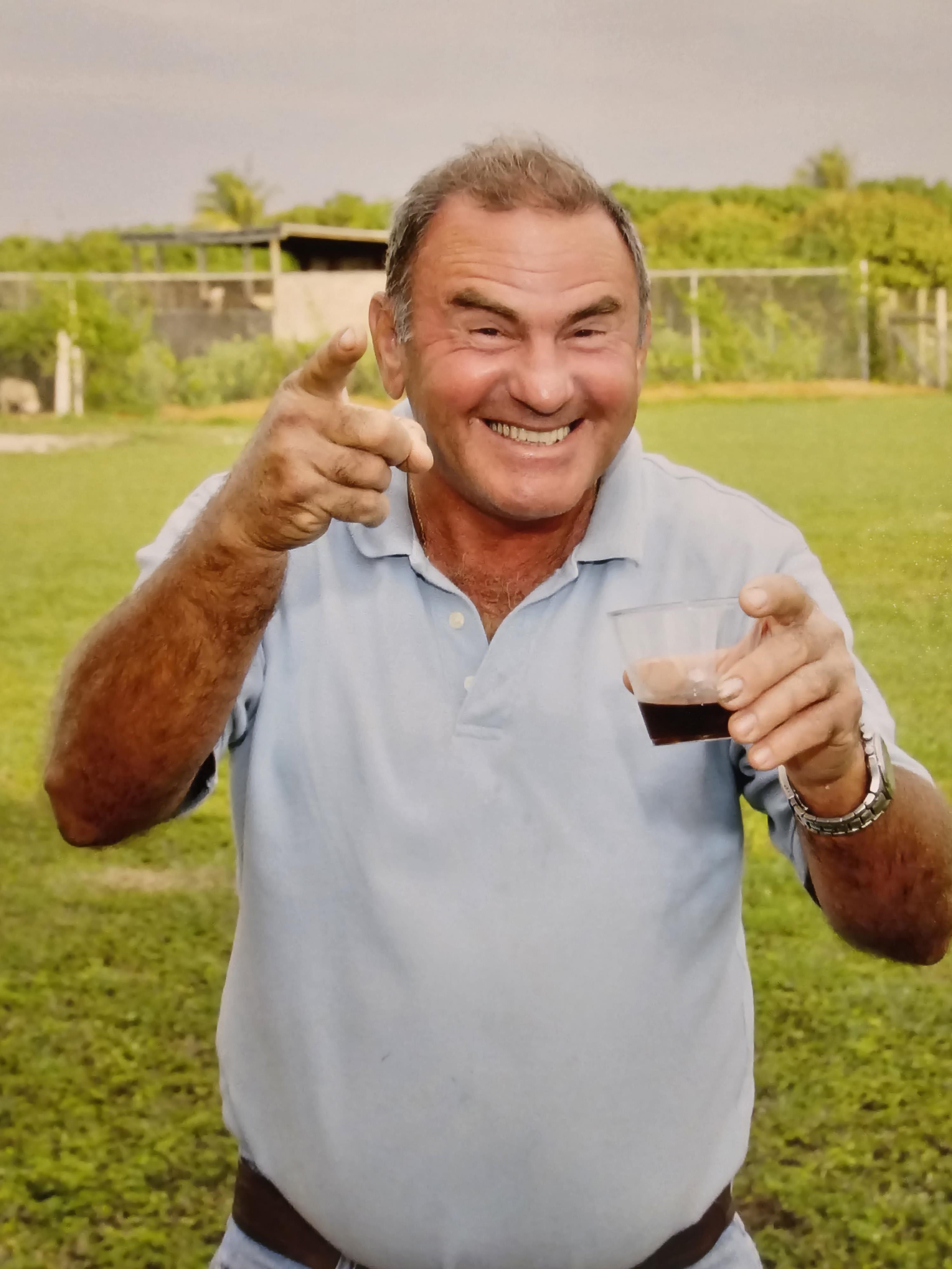 A man stands in a green field, smiling widely and holding a drink while pointing at the camera.