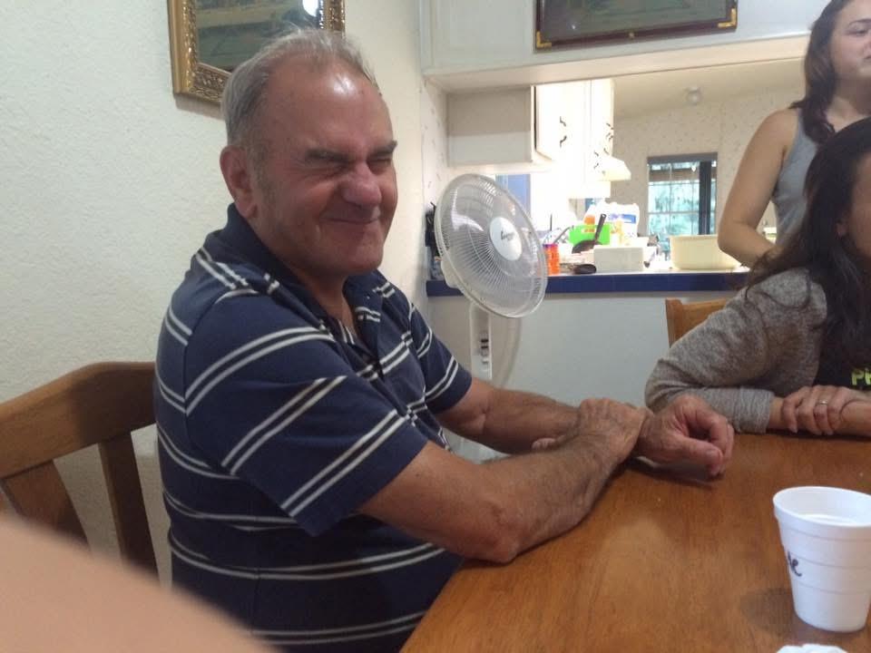Elderly man smiles and makes a funny face while sitting at a table with friends and a fan.