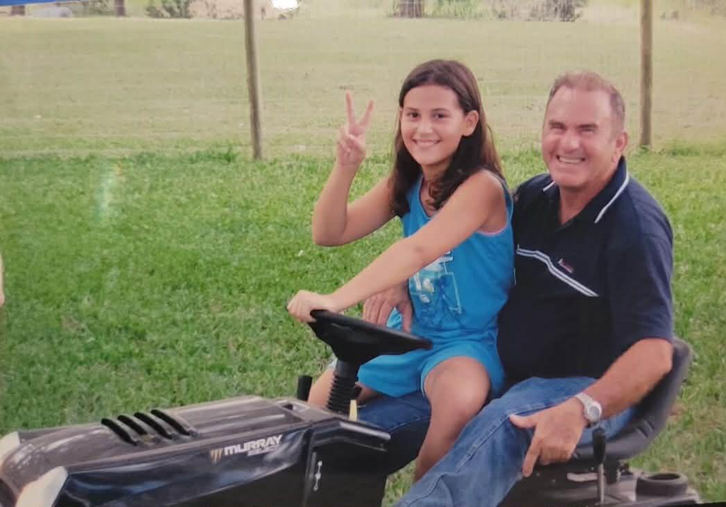 A young girl sits on a lawn mower with an adult man in a backyard. They smile and have fun.