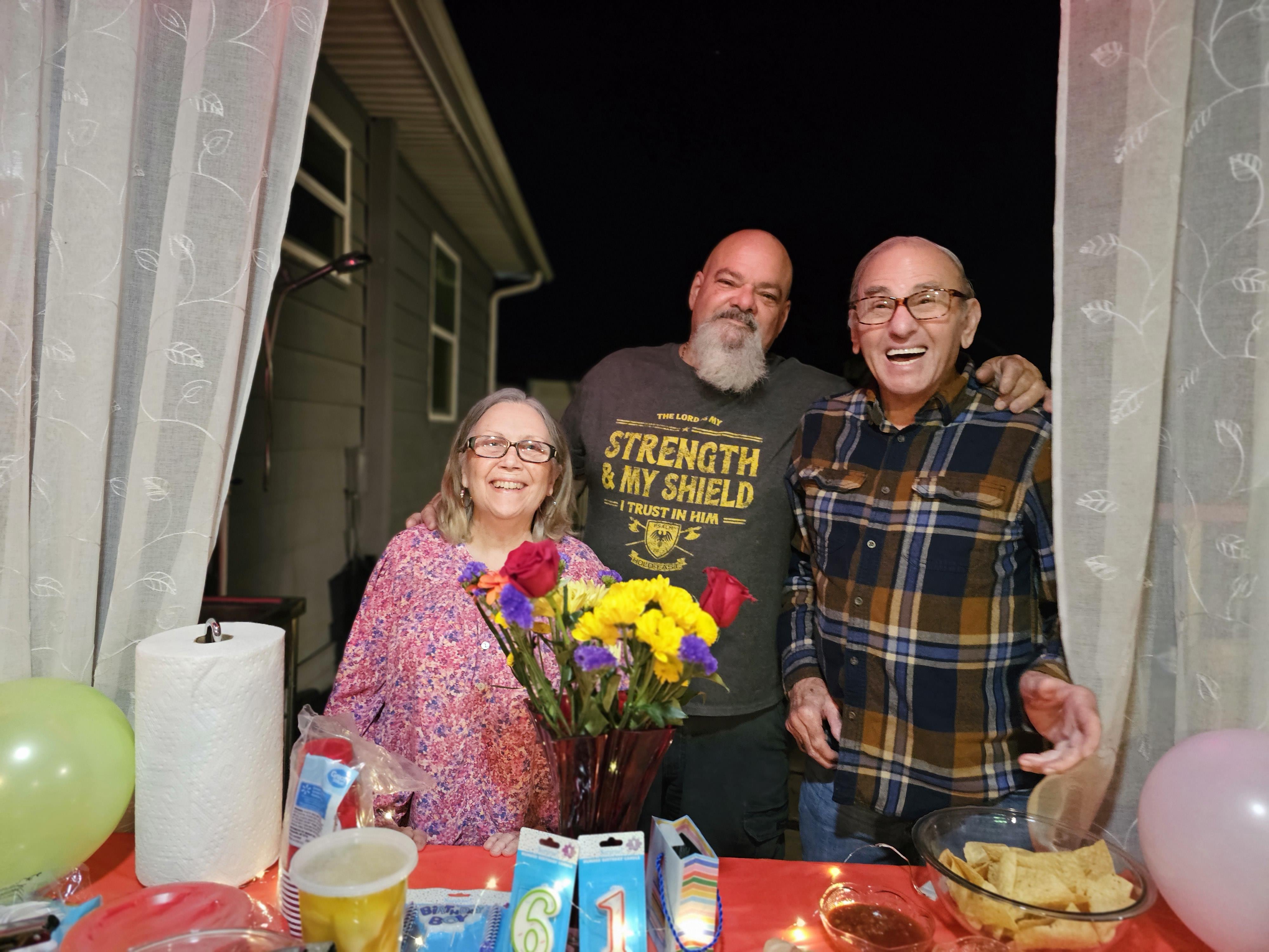 Friends gather on a porch at night, smiling and enjoying food while flowers decorate the table.