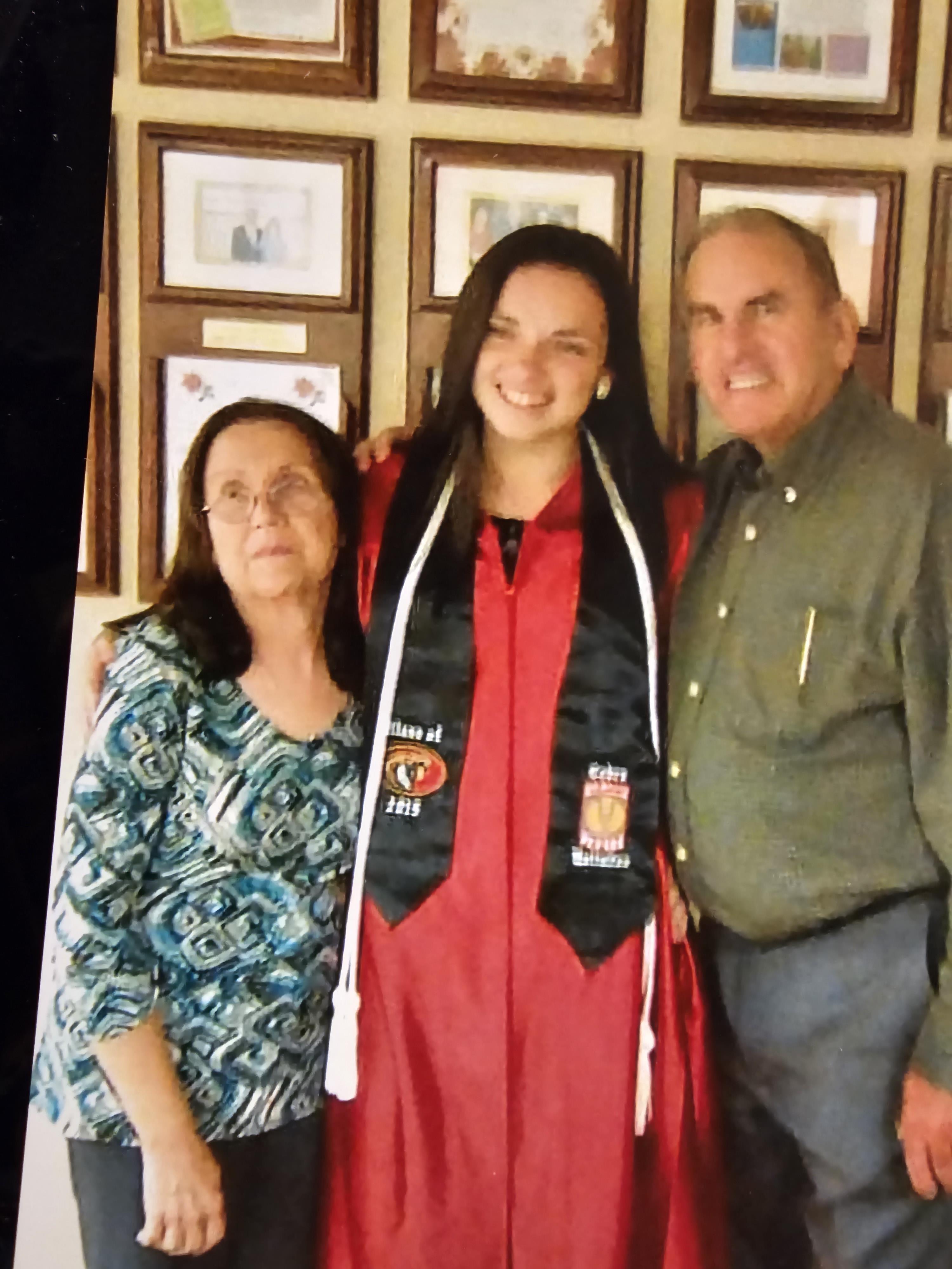 A young woman in a graduation gown stands proudly with her parents at home.