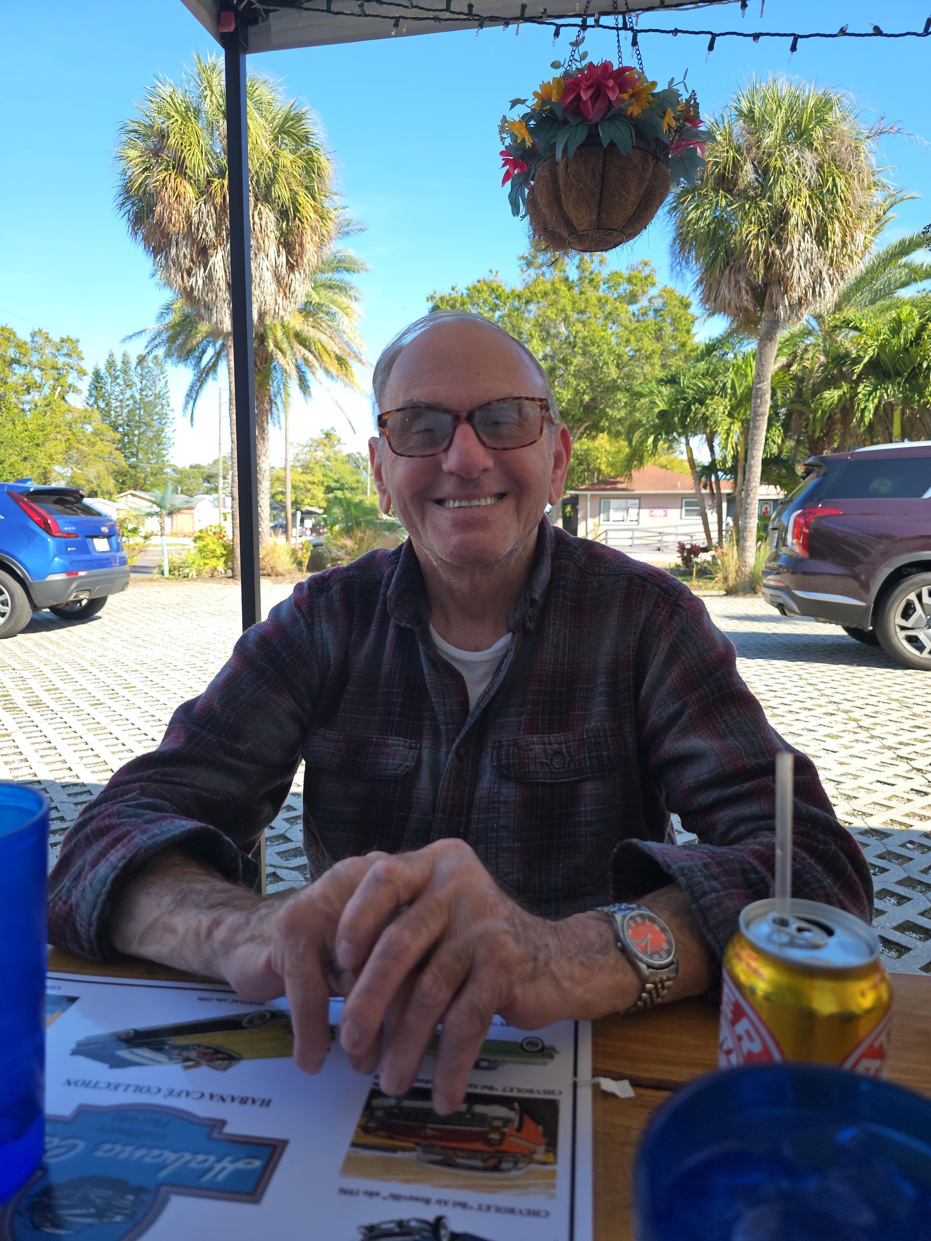 Man smiles while sitting at a table outside, enjoying his drink in a sunny location.
